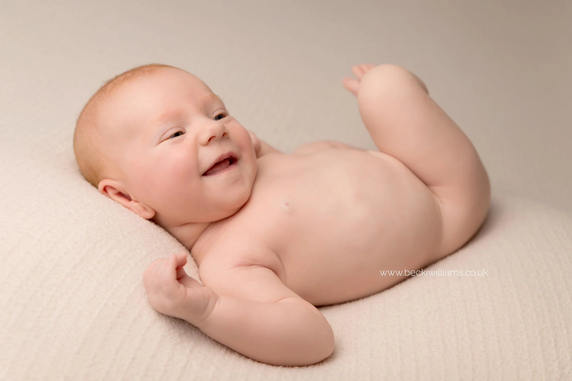 newborn baby girl laying on her back on a cream blanket, smiling, for her newborn photo shoot in hertfordshire