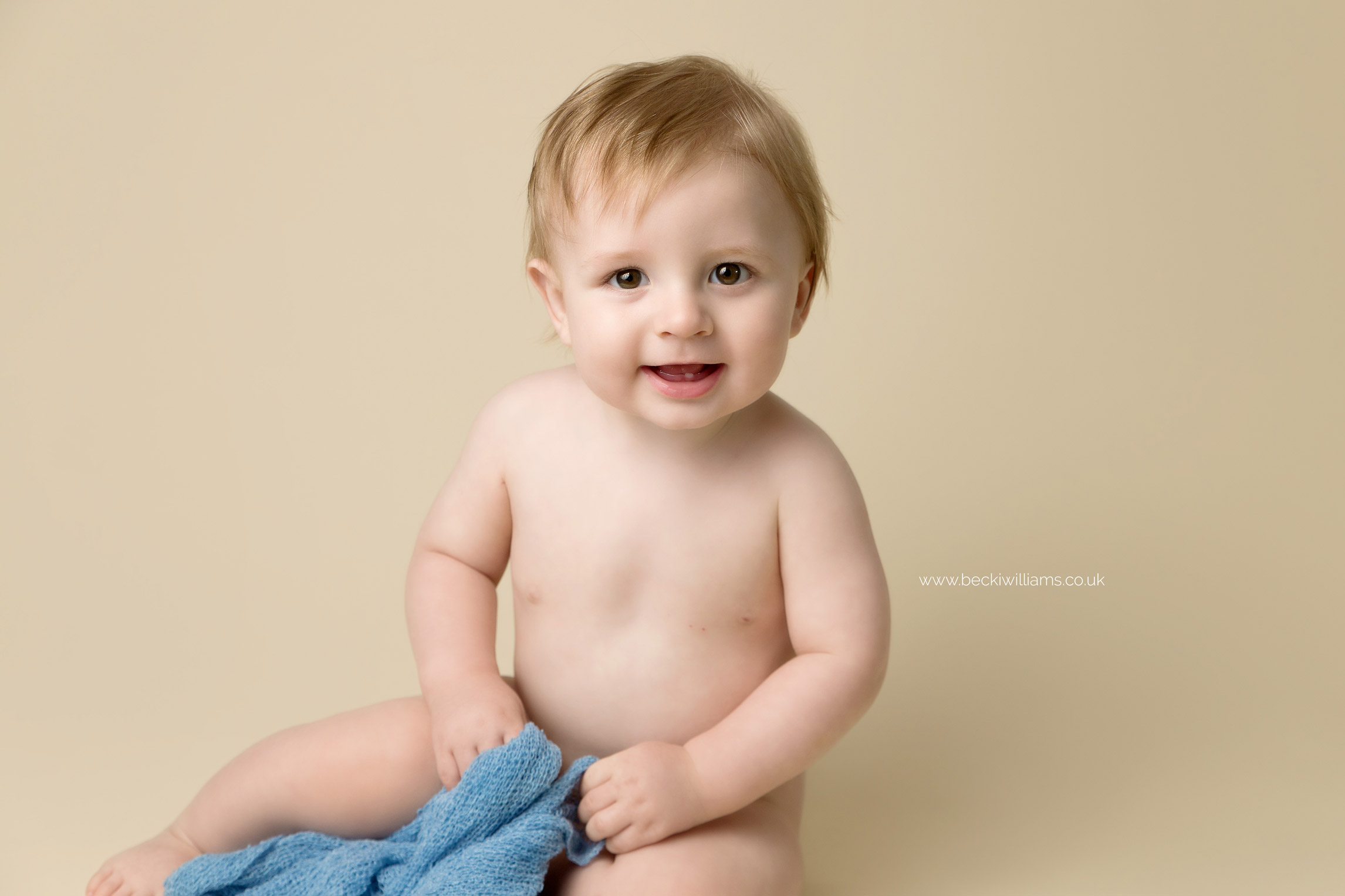 7 month old baby boy smiling, sitting on a cream backdrop at his baby photo shoot in Buckinghamshire