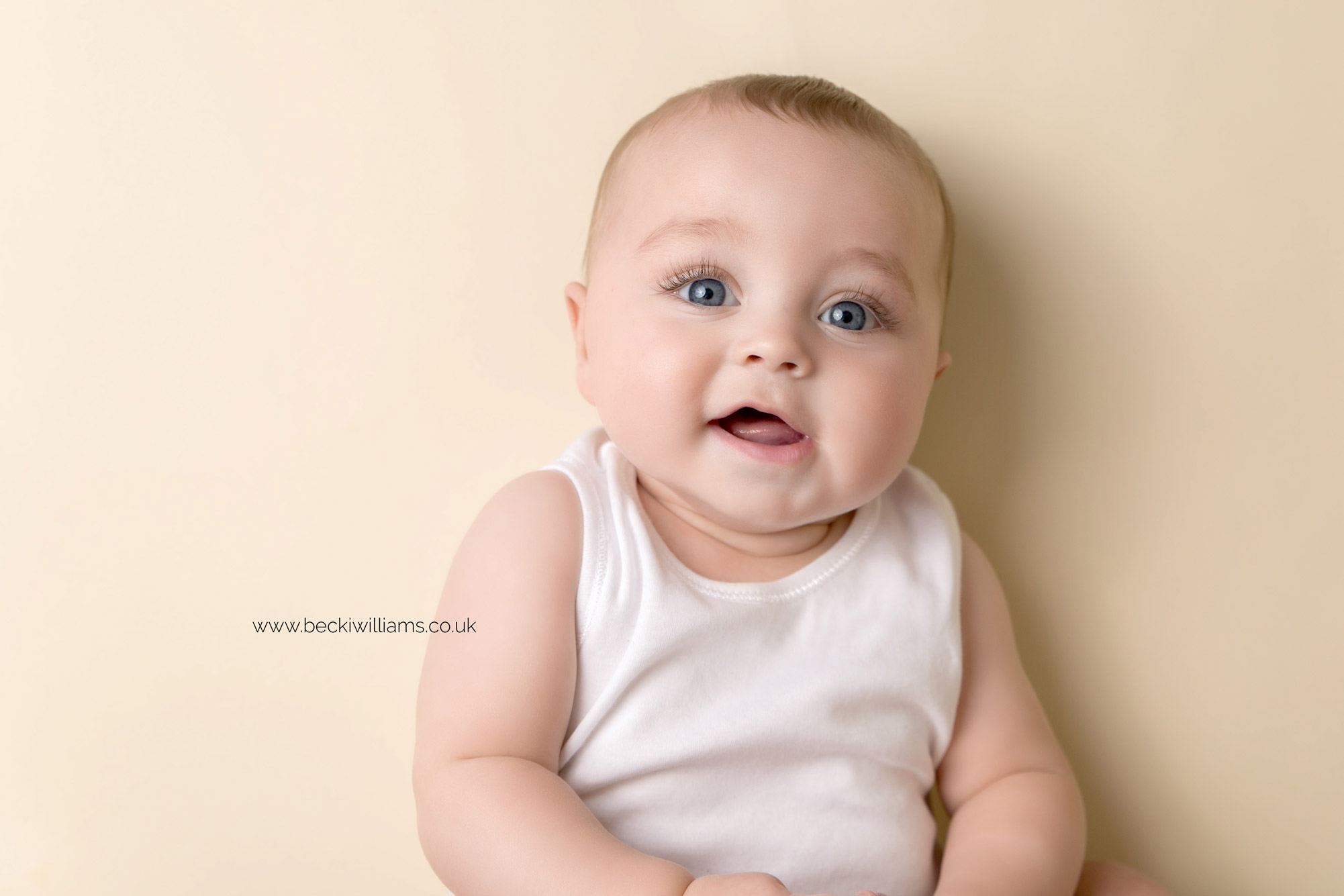 5 month old baby boy lays on the floor, looking up at the camera smiling.  He is wearing a white vest