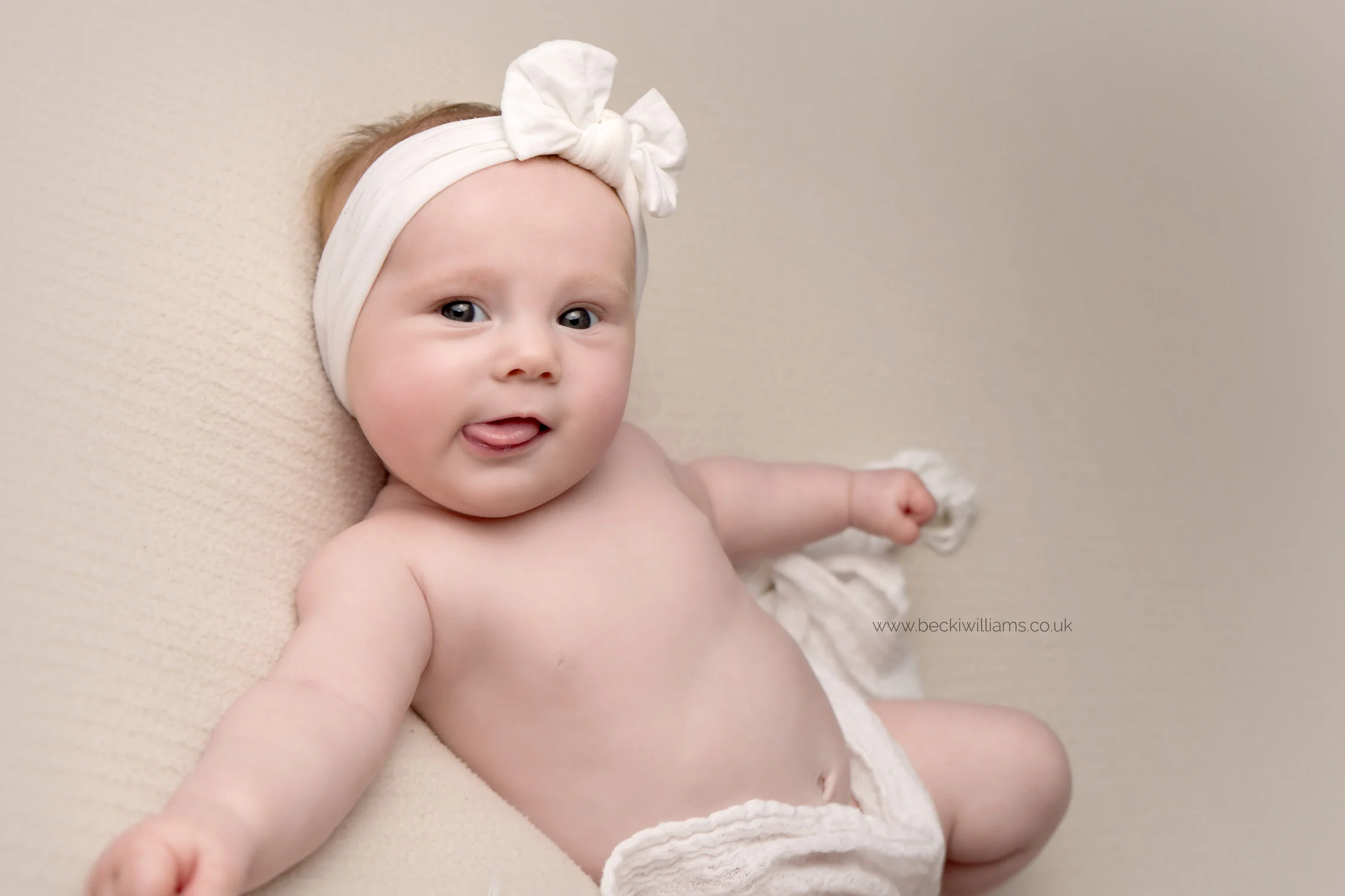 2 month old baby girl lays on her back, smiling with her tongue sticking out at her baby photo shoot in hemel hempstead