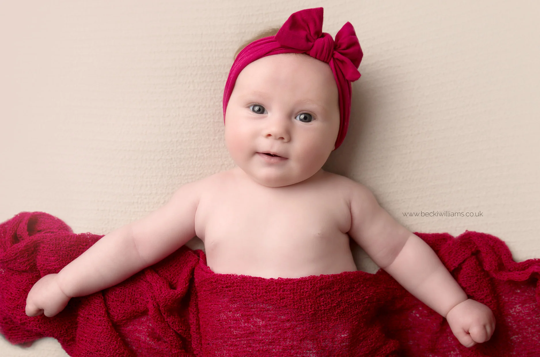 2 month old baby girl wearing a red headband, covered in a red blanket at her photo shoot in hemel hempstead