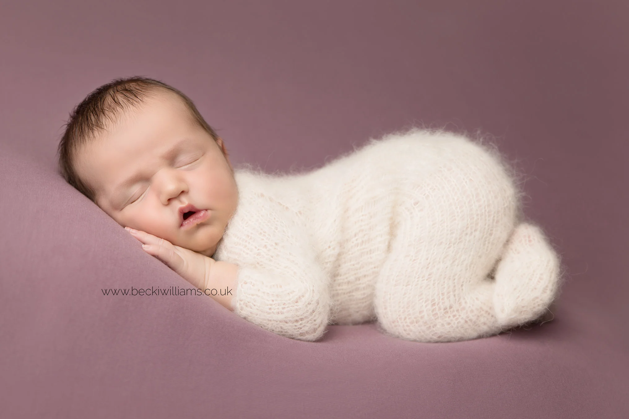 newborn girl portrait of a baby wearing a white fluffy outfit, curled up sleeping on her front, laying on a pink blanket at her newborn photo shoot in hemel hempstead