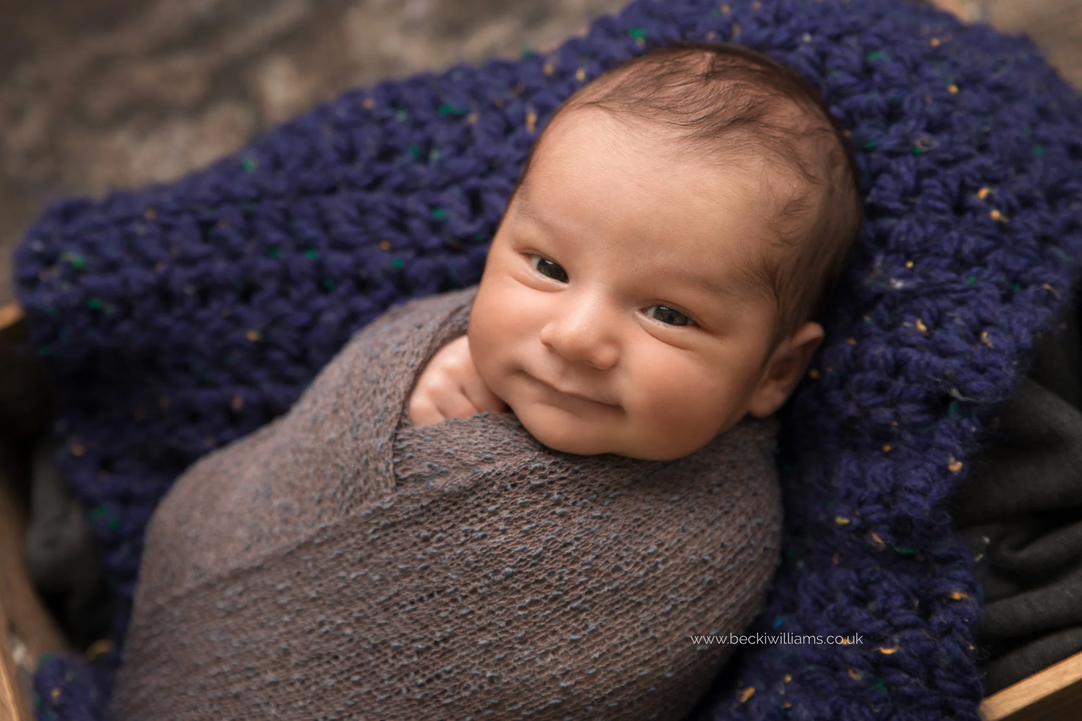 close up of wrapped, smiling newborn baby in a crate for his newborn photo shoot in hemel hempstead