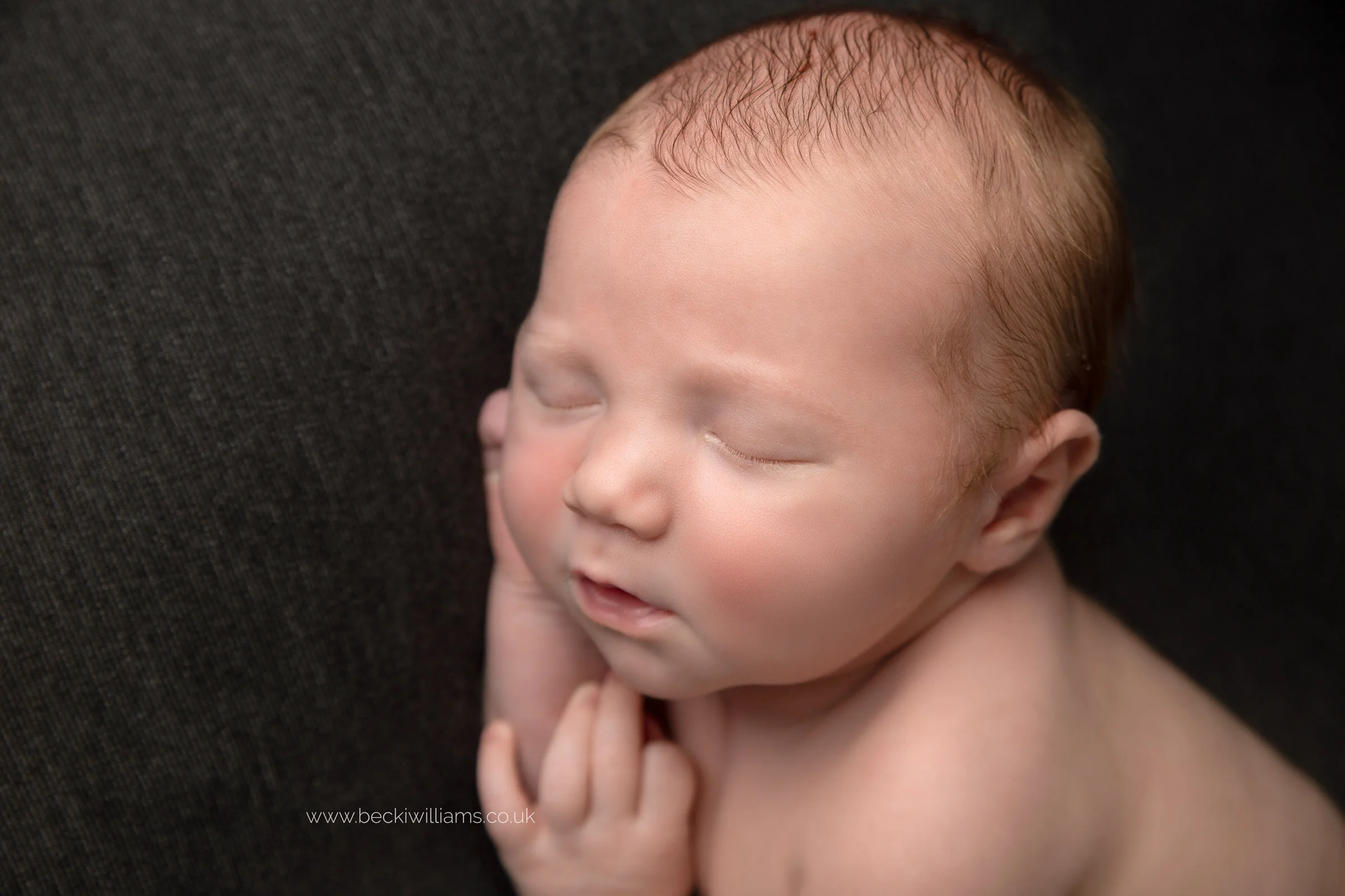 Newborn baby boy lays asleep on his side on a dark grey blanket at his newborn shoot in Hemel Hempstead.