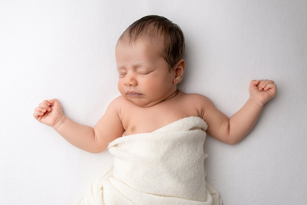 Newborn baby lays asleep on a white blanket, wrapped in a white wrap