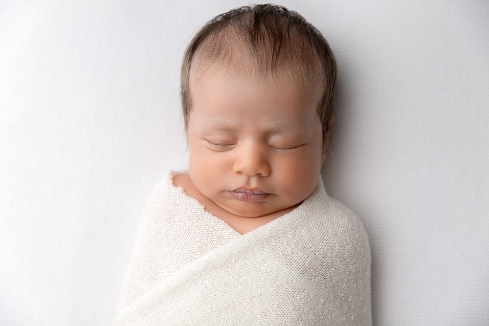 Newborn baby lays asleep on a white blanket, wrapped in a white wrap