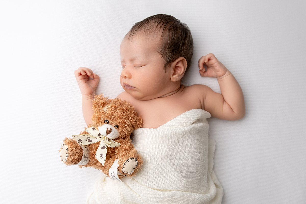 Newborn baby lays asleep on a white blanket, wrapped in a white wrap with a brown bear