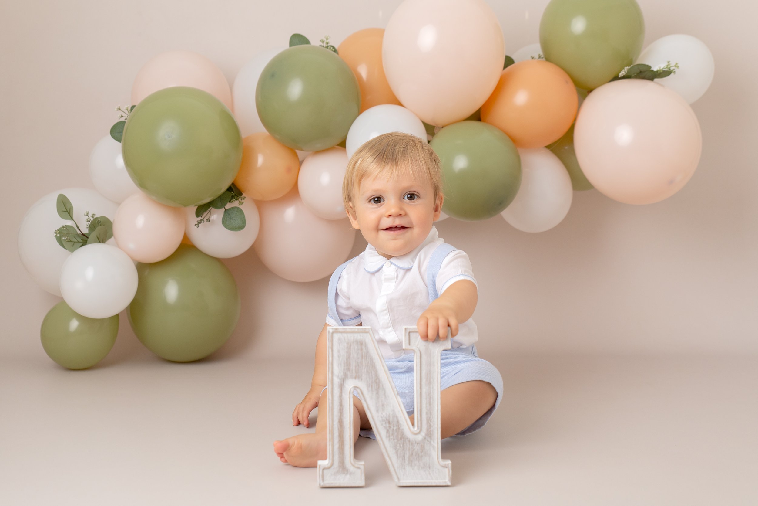 1 year old boy at his birthday photo shoot.  He’s sitting on the floor in front of a green, peach and white balloon arch.  He holds the letter N. 