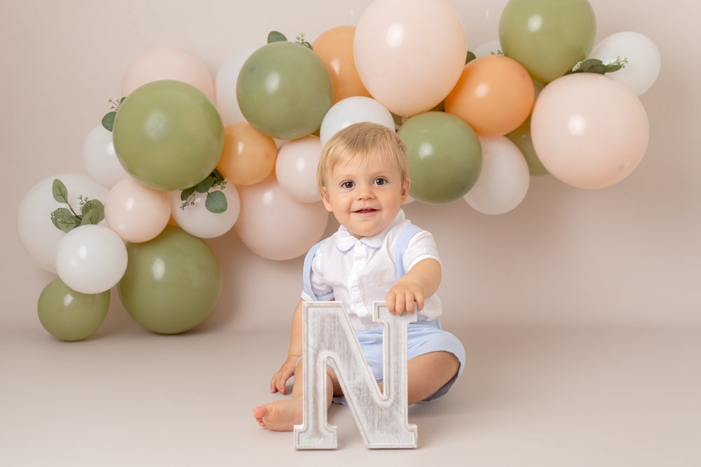  1 year old boy at his birthday photo shoot.  He’s sitting on the floor in front of a green, peach and white balloon arch.  He holds the letter N. 