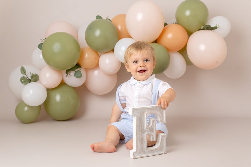  1 year old boy at his birthday photo shoot.  He’s sitting on the floor in front of a green, peach and white balloon arch.  He holds the letter E. 