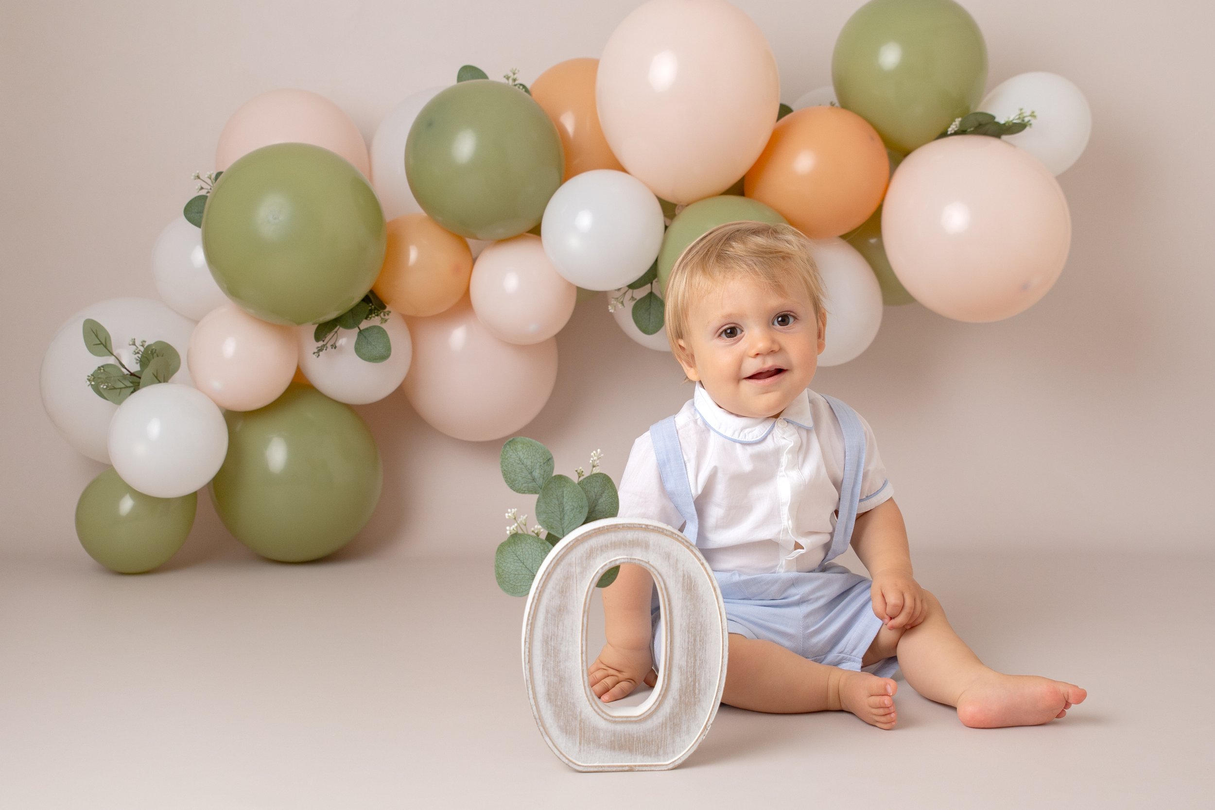  1 year old boy at his birthday photo shoot.  He’s sitting on the floor in front of a green, peach and white balloon arch.  He holds the letter O. 
