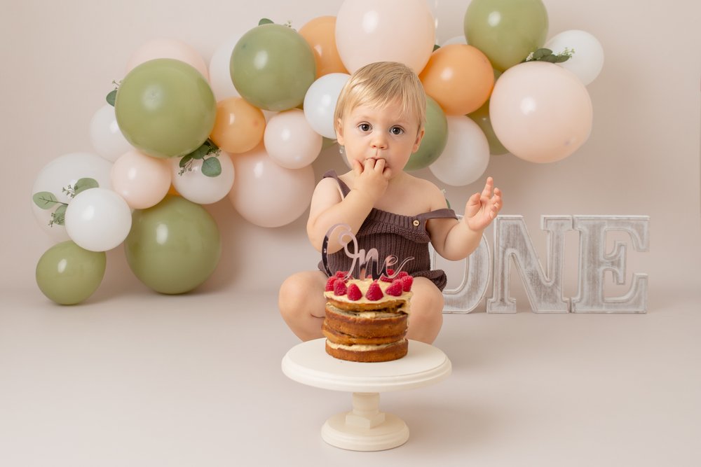  1 year old at his cake smash photo shoot in Milton Keynes.  He is sitting in front of a green, peach and white balloon garland and the letters ONE.  There is a cake in front of him.  