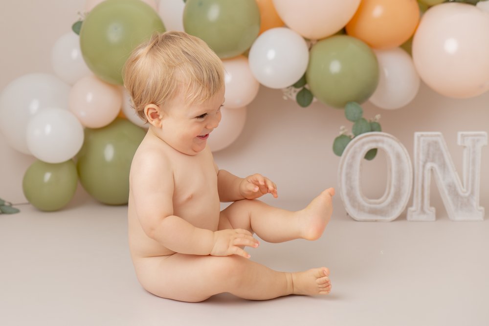  1 year old at his cake smash photo shoot in Milton Keynes.  He is sitting in front of a green, peach and white balloon garland and the letters ONE.  