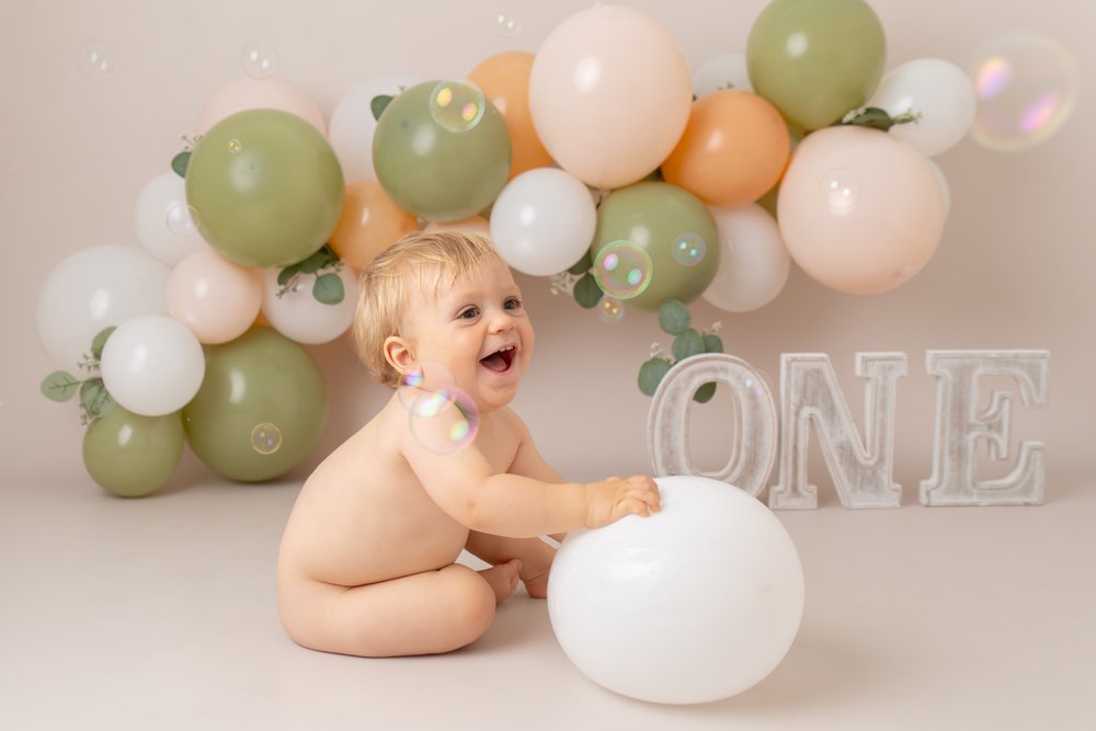  1 year old at his cake smash photo shoot in Milton Keynes.  He is sitting in front of a green, peach and white balloon garland and the letters ONE.  He’s holding a white balloon. 