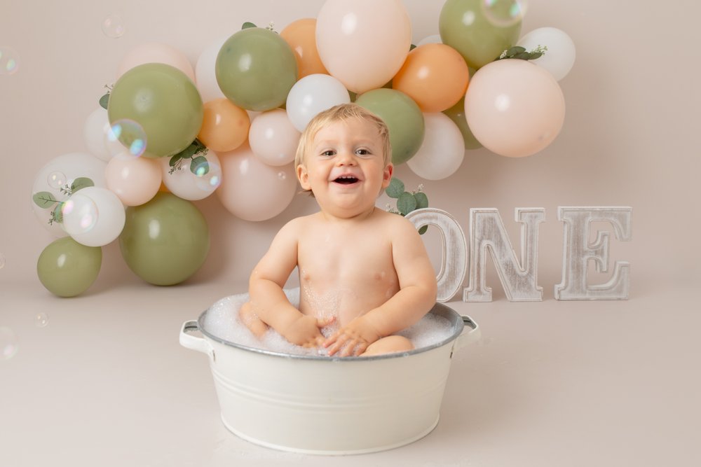 1 year old at his cake smash photo shoot in Milton Keynes.  He is sitting in a tin bath front of a green, peach and white balloon garland and the letters ONE.  