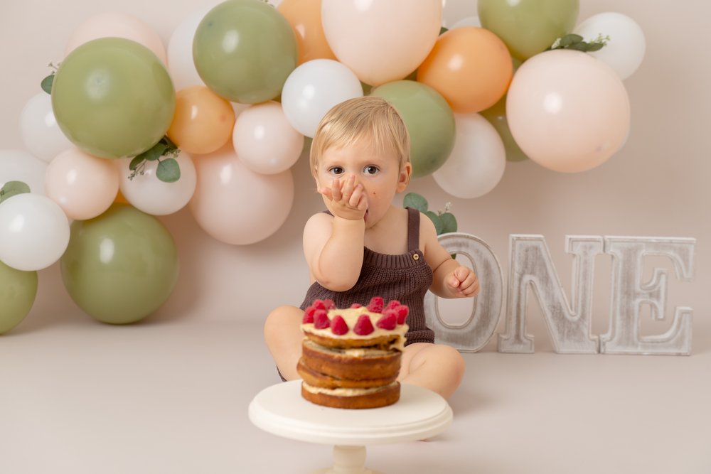  1 year old at his cake smash photo shoot in Milton Keynes.  He is sitting in front of a green, peach and white balloon garland and the letters ONE.  There is a cake in front of him.  
