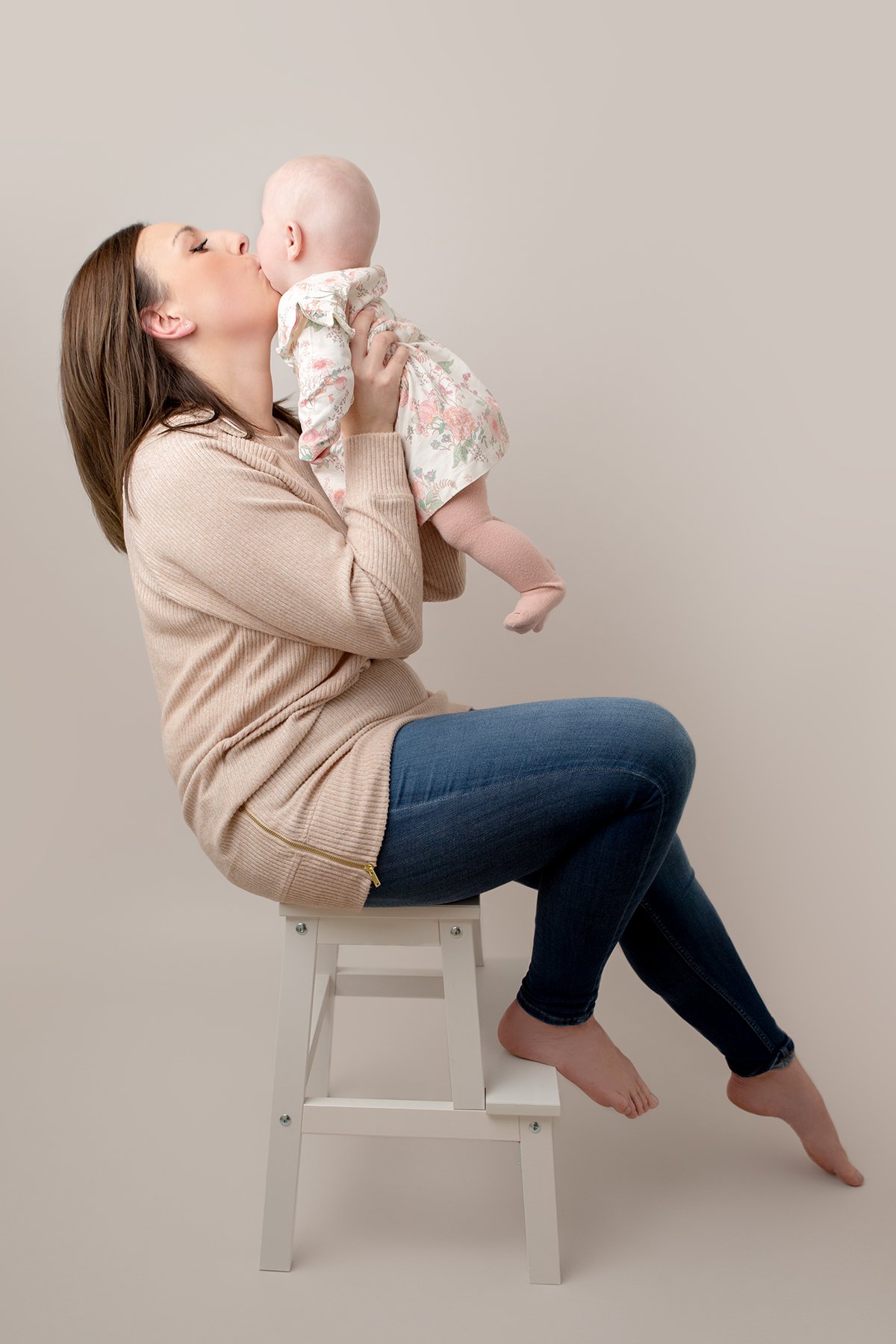Mum sitting on a stool holding up and kissing her 3 month old baby girl at their professional photo shoot
