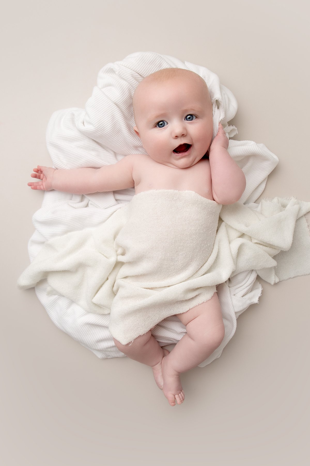 Professional baby photo of a young baby laying on their back, covered in a white blanket