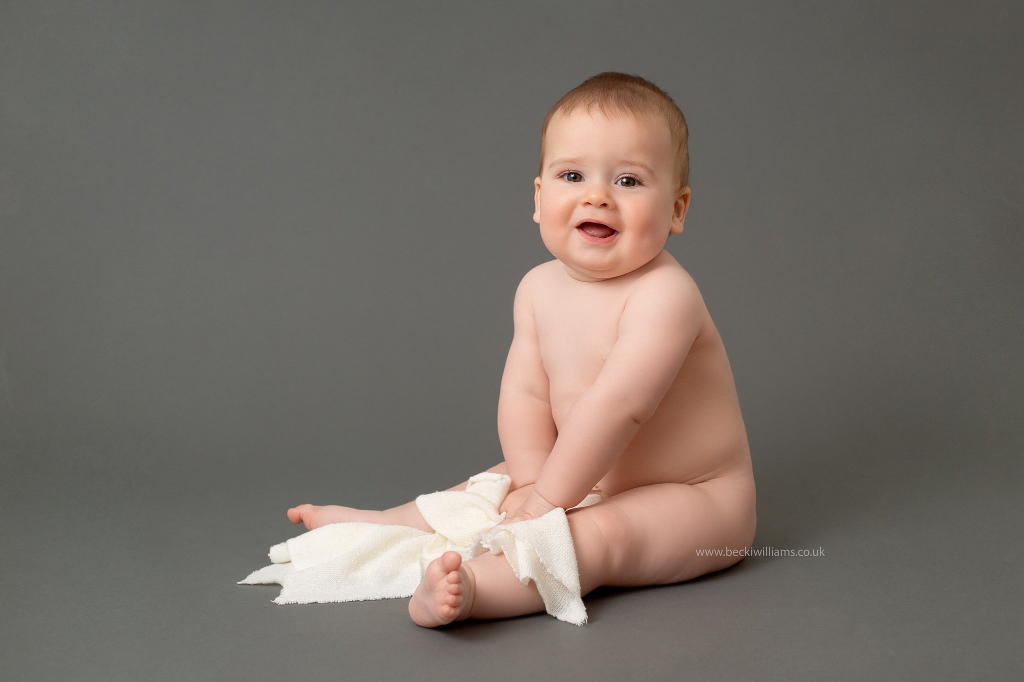 9 month old boy sits on a dark grey backdrop with a white blanket at his baby photo shoot