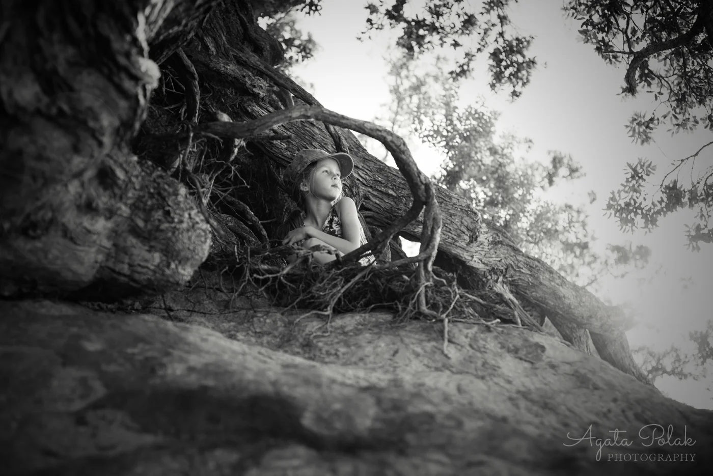 A young girl with a hat sitting in a tree hollow, looking contemplative, surrounded by large tree roots and branches in a black and white photograph.