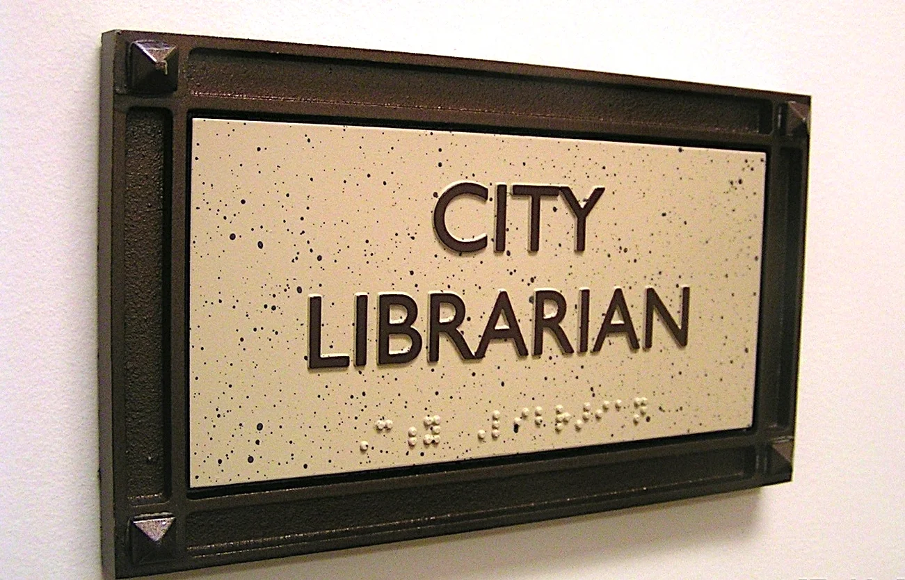  ADA Room ID - Photopolymer plaque with raised silkscreened text, tactile braille, painted faux stone background finish, cast aluminum decorative sign frame with painted finish. 