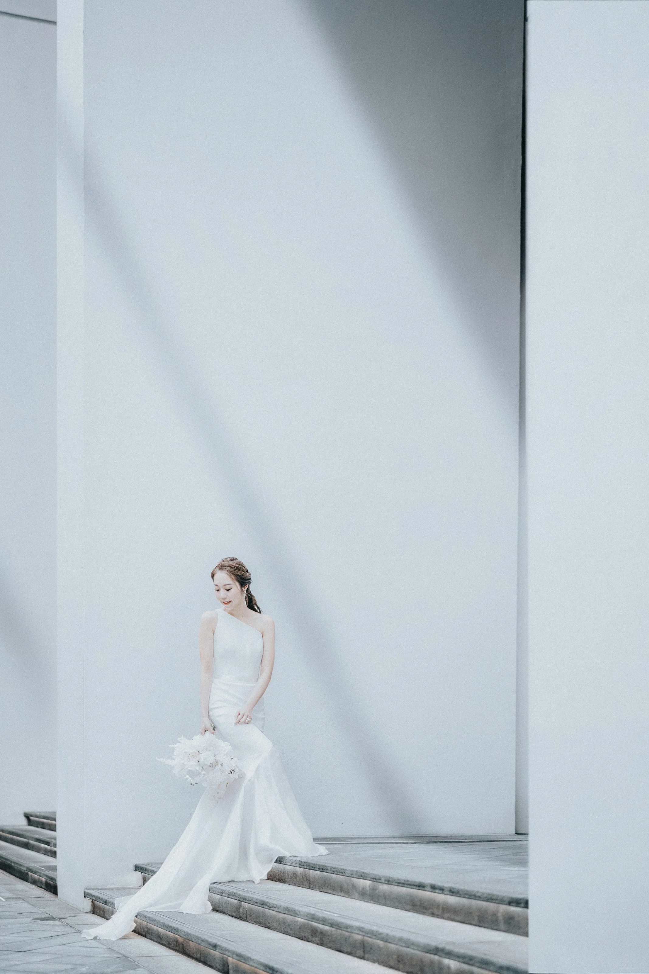 A woman in a white wedding dress holding a bouquet standing on modern steps outdoors with minimalist white walls and soft diagonal shadows.