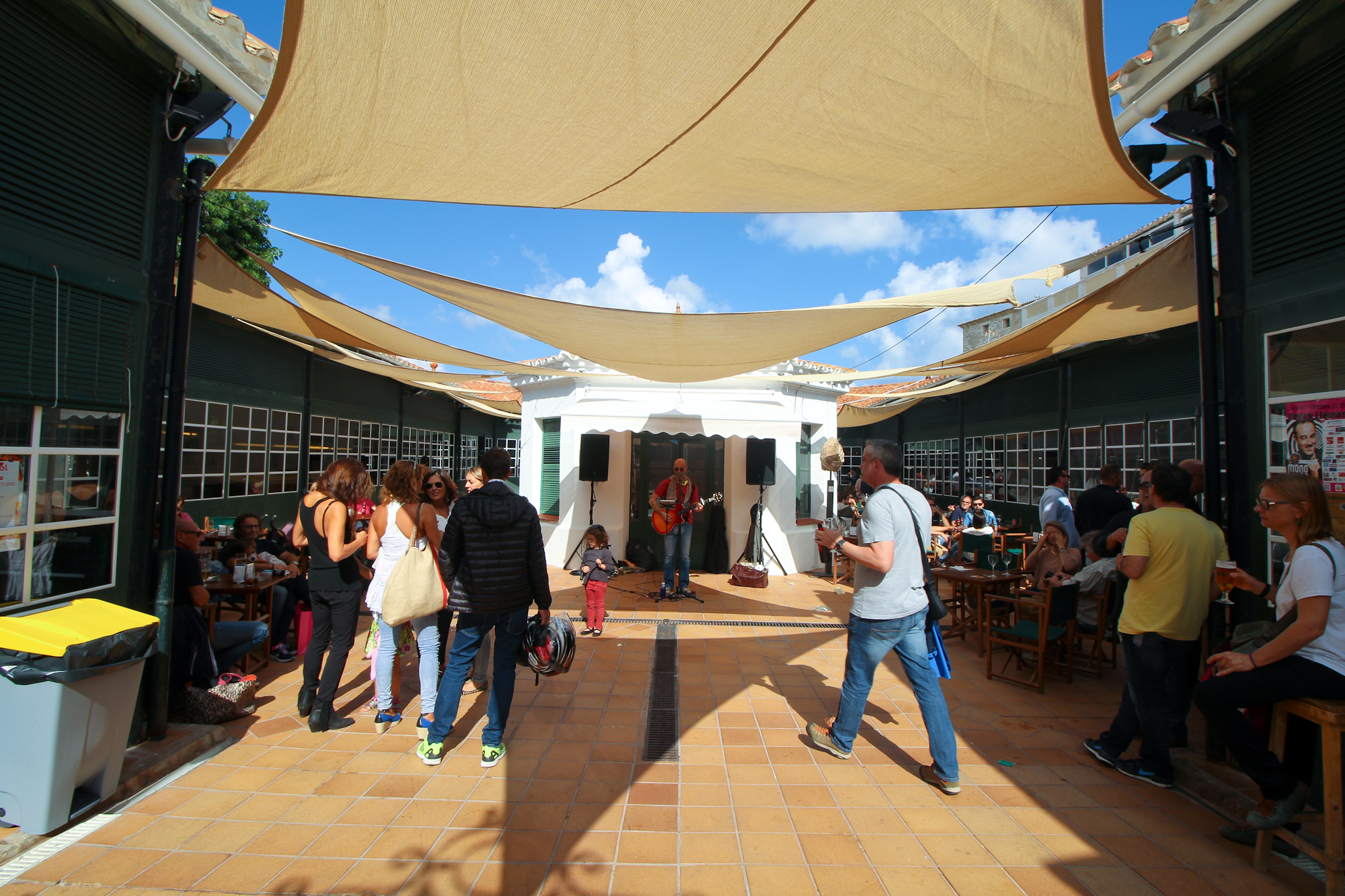 You enter into a large open area (atrium?). As you can see there's a man with a guitar, tables all around the side with merry people, and you can enter the surrounding building on either side to grab tapas and drinks.