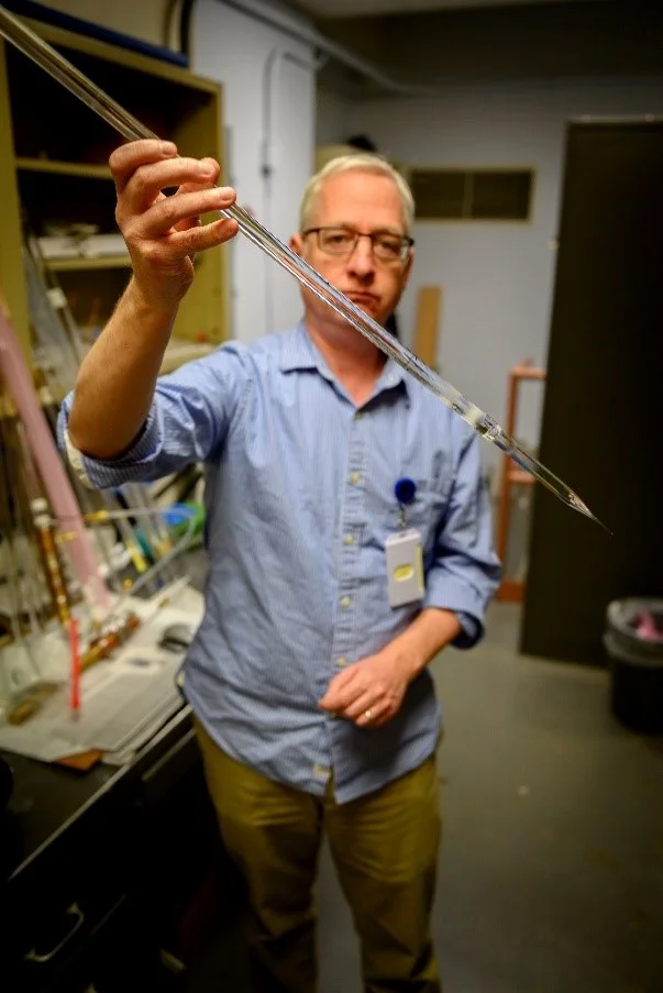 Colin Baker, U.S. Naval Research Laboratory, holds a silica glass rod (optical preform) that will be pulled into an optical fiber suitable for production of an eye safer laser at U.S. NRL, DC on March 26, 2019. (U.S. Navy photo by Jonathan Steffen)