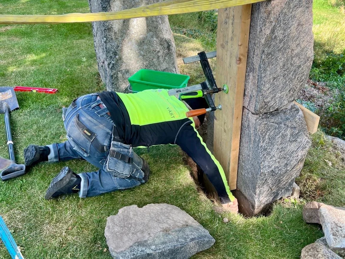  Nästa steg är att slå ner rejäla kilstenar framför runstenen så den fixeras i lodrät position. För att dessa ska få utrymme gräver vi undan jordmassor. Notera storleken på kilstenarna, de går djupt ner i marken. 