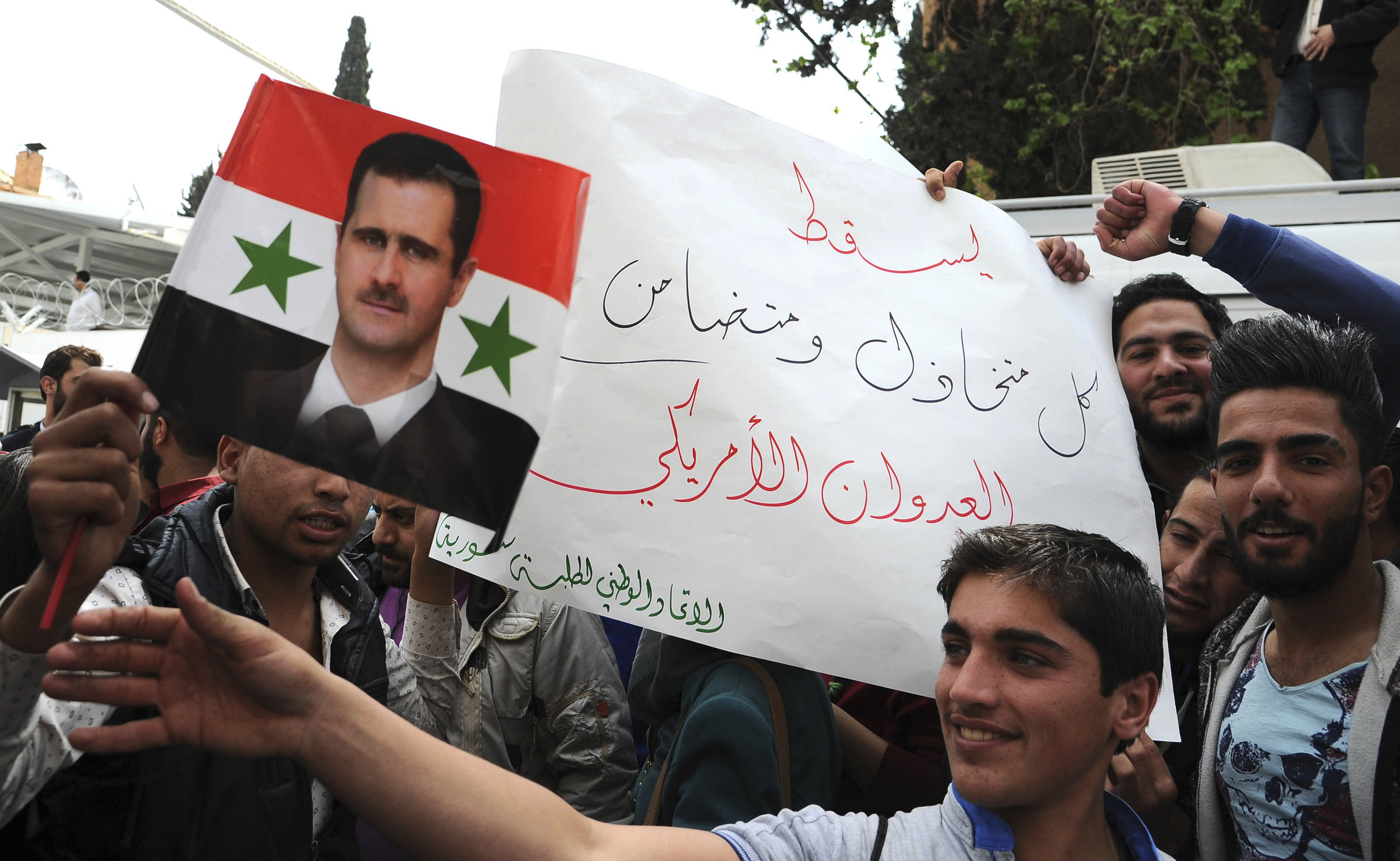 In this photo released by the Syrian official news agency SANA, pro-government protesters hold a portrait of President Bashar Assad and a placard that reads, "Down with everyone who cooperated and supported the American aggression," during a protest in front of the the United Nations building, in Damascus, Syria, Tuesday, April 11, 2017. (SANA via AP)