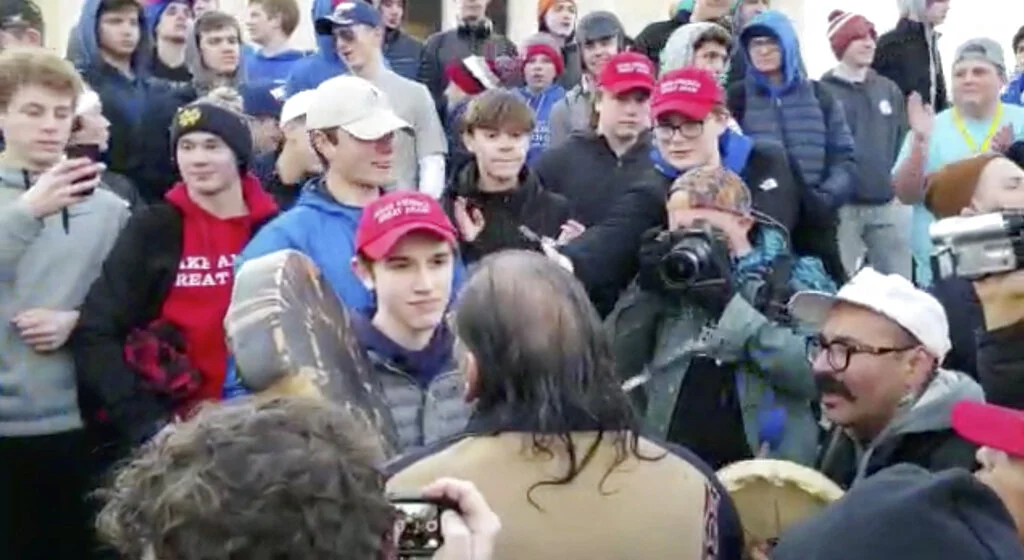 In this Friday, Jan. 18, 2019 image made from video provided by the Survival Media Agency, a teenager wearing a "Make America Great Again" hat, center left, stands in front of an elderly Native American singing and playing a drum in Washington. The Roman Catholic Diocese of Covington in Kentucky is looking into this and other videos that show youths, possibly from the diocese's all-male Covington Catholic High School, mocking Native Americans at a rally in Washington. (Survival Media Agency via AP)