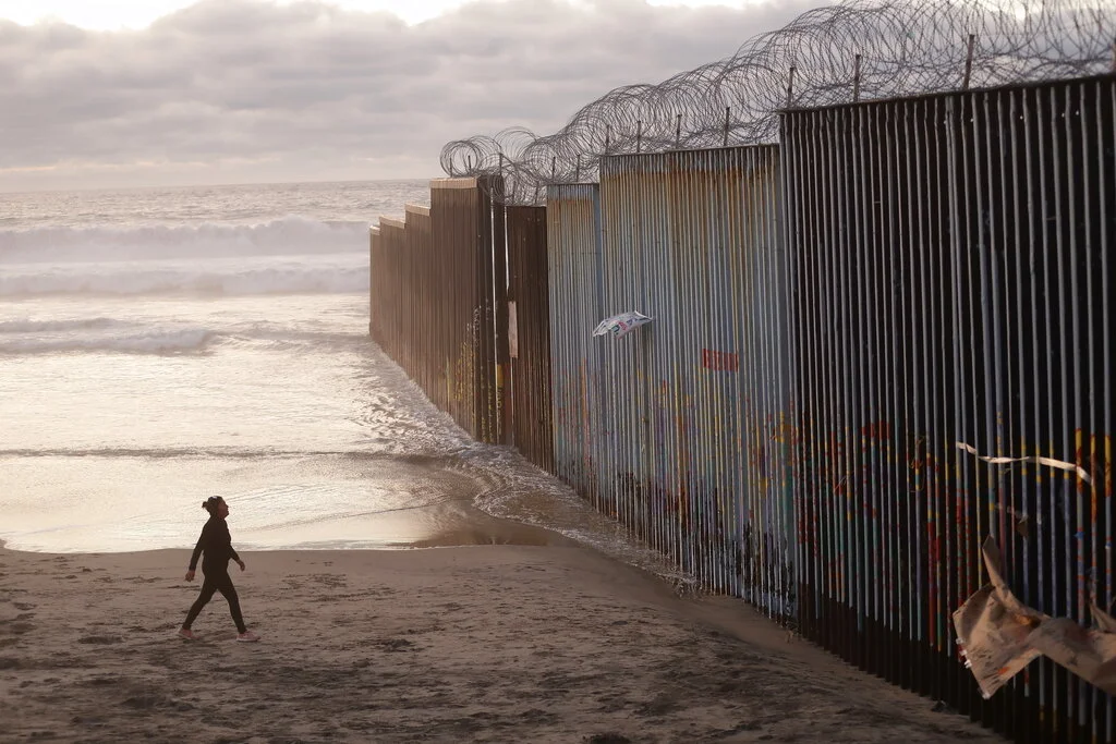 FILE - In this Jan. 9, 2019 file photo, a woman walks on the beach next to the border wall topped with razor wire in Tijuana, Mexico. A group of former U.S. national security officials is set to release a statement on Monday, Feb. 25, arguing that there is no justification for President Donald Trump to use a national emergency declaration to fund a wall along the U.S.-Mexico border. (AP Photo/Gregory Bull, File)