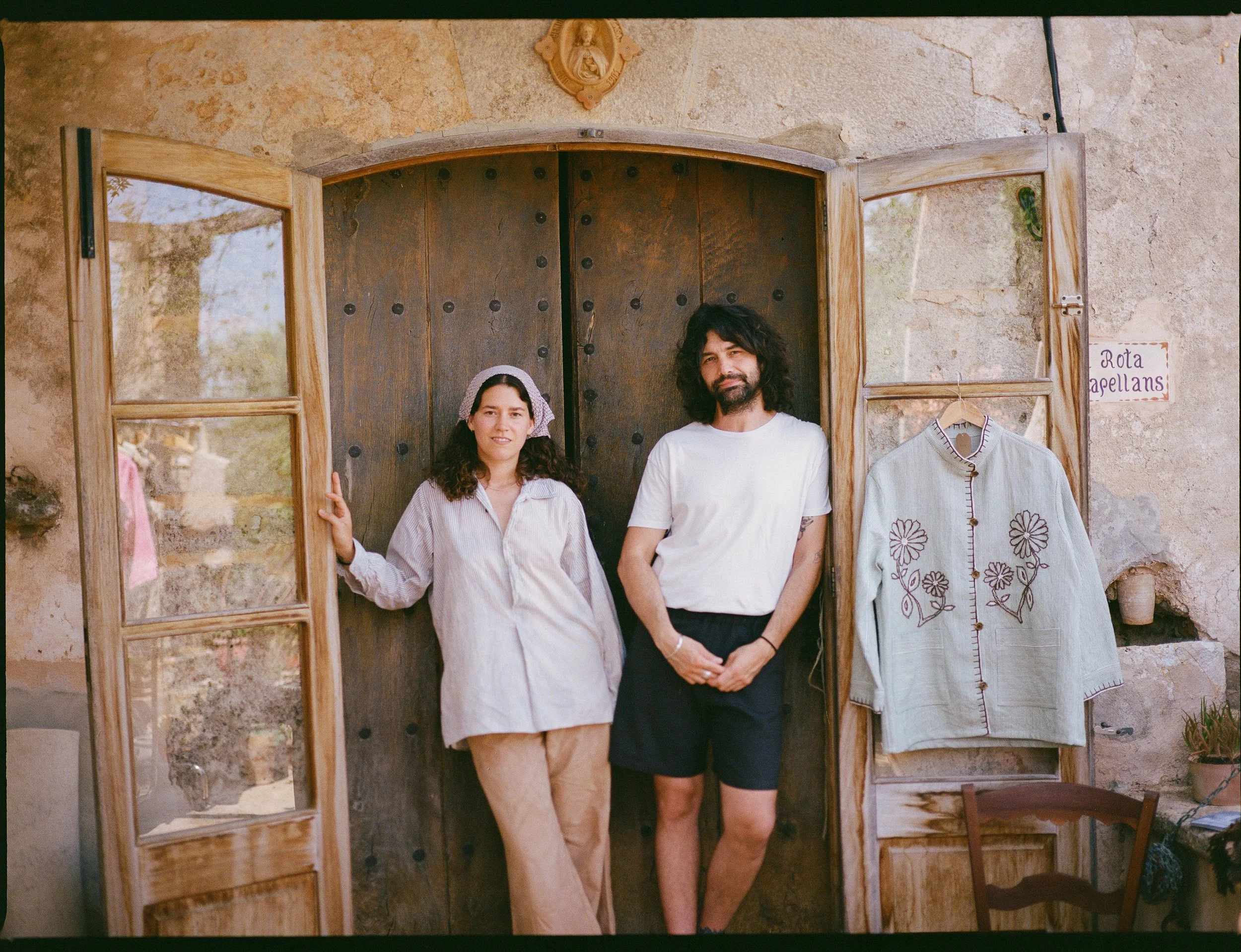 A woman and a man standing in front of a rustic wooden door with open window panels, inside a stone-walled room. The woman is wearing a light-colored shirt and beige pants, with a headscarf. The man is wearing a white t-shirt and black shorts. To the right, a light gray embroidered jacket hangs on the wall, and a small sign reads 'Rota Capellans.'
