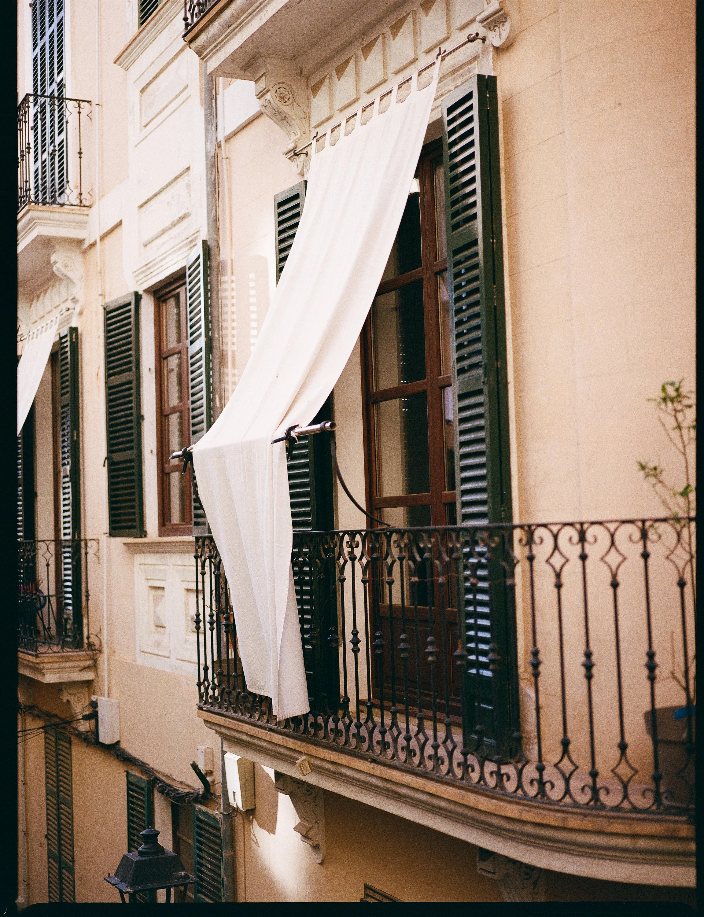 A beige building with green shutters and a small balcony, featuring a white curtain blowing in the wind. Classic architecture of Palma de Mallorca, Spain.