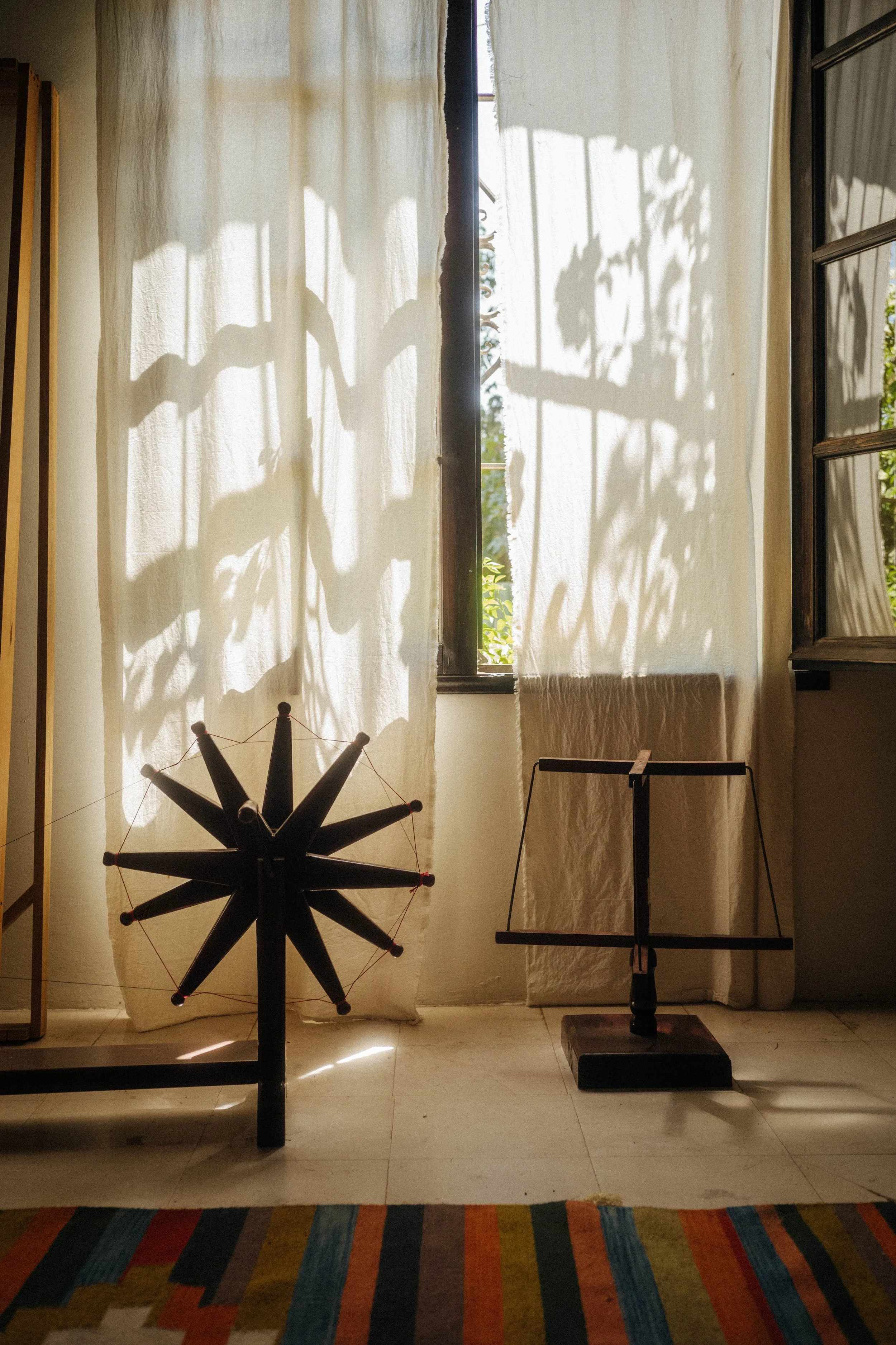 Sunlight filters through a window, casting shadows of leaves and branches on white curtains inside a room. There is a spinning wheel on the floor.