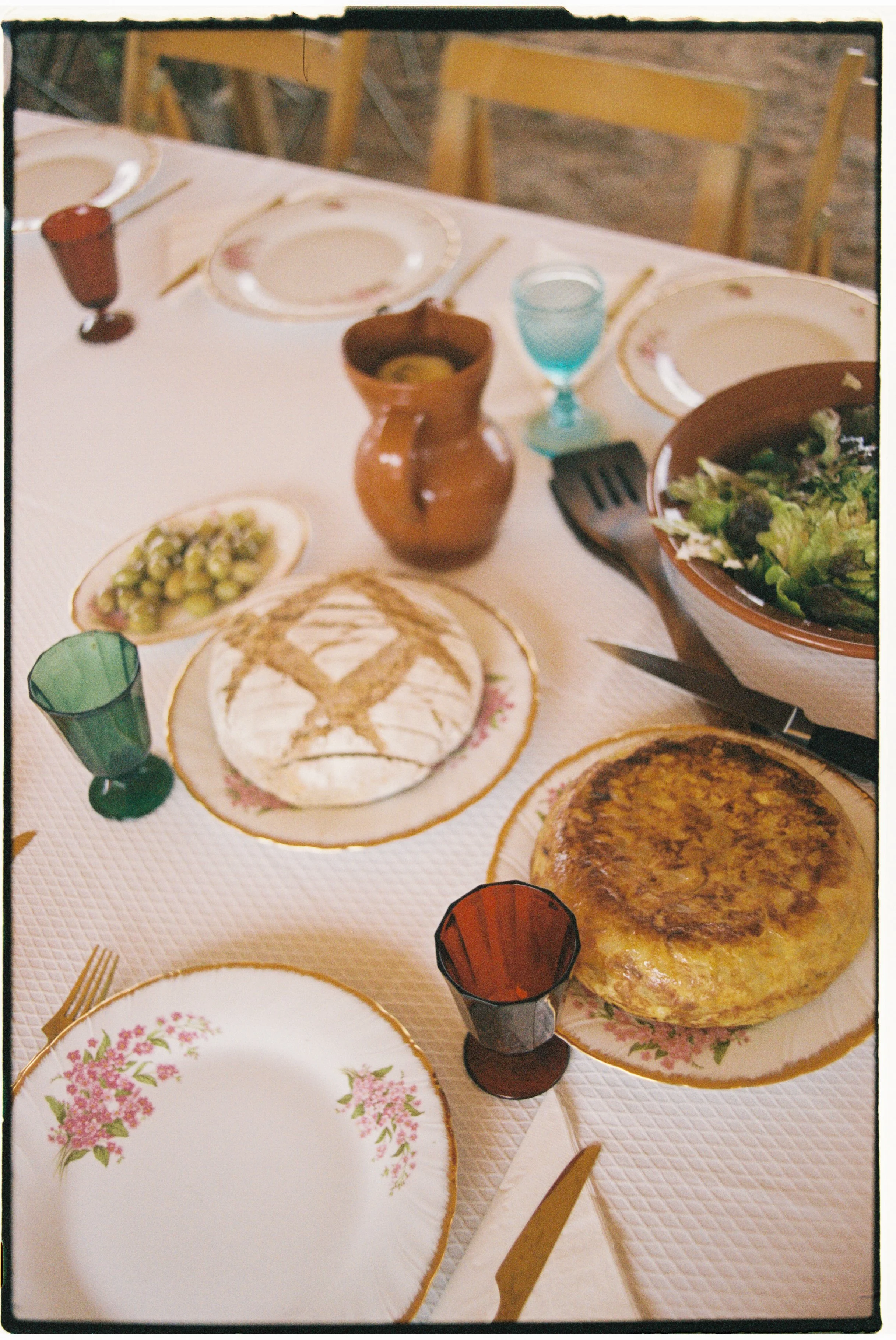 A table set for a meal with plates, glasses, bread, a bowl of salad, a tortilla, cheese, cutlery, and a small pitcher on a white tablecloth. At the Accidente Con Flores textile studio.