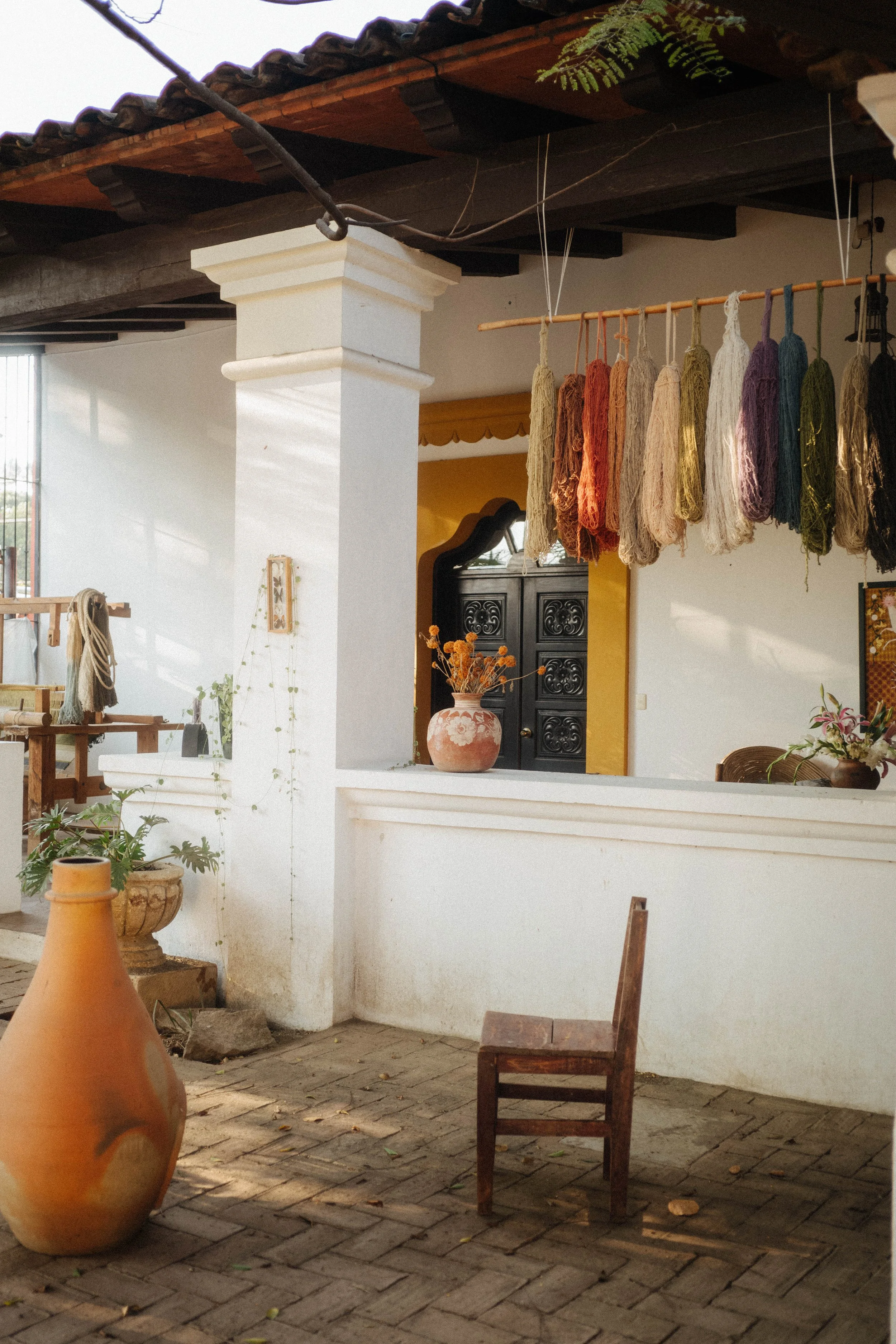 Interior of a cozy yarn shop with colorful skeins hanging from the ceiling, potted plants, vases, a wooden chair, and shelves with yarn and craft supplies.