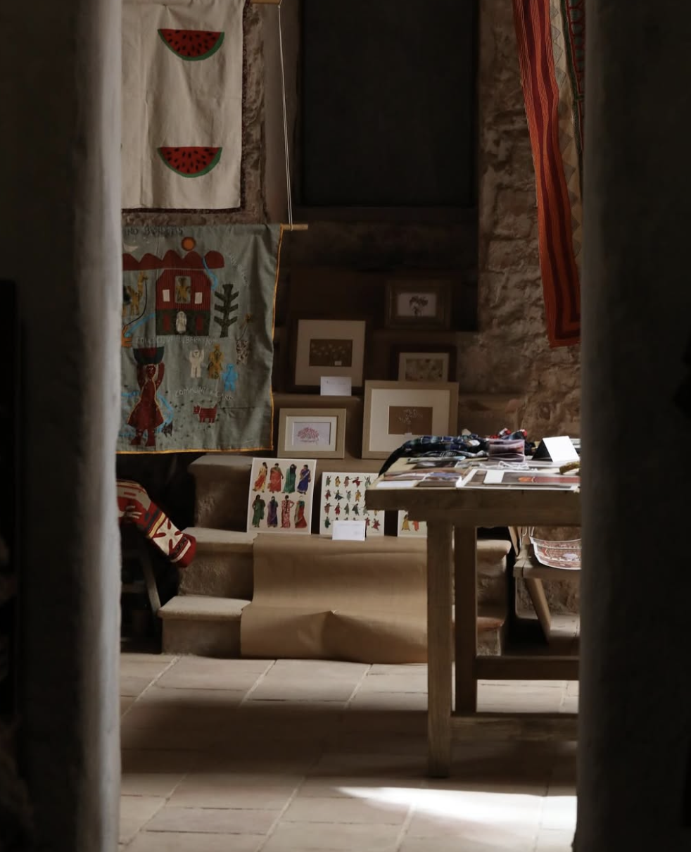 View of an art display inside a stone-walled room, including framed artwork, fabric quilts, and embroidery pieces, with a table holding printed materials and a woven chair.