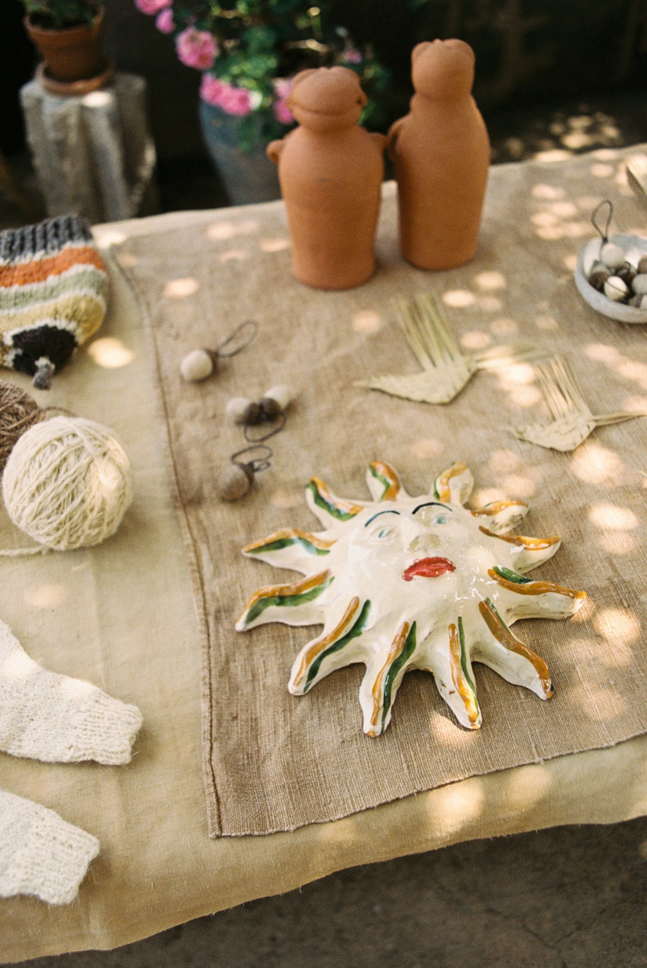 Decorative sun-shaped ceramic plate with a face, positioned on a beige tablecloth among various pottery and handmade crafts at the Accidente Con Flores studio in Mallorca.
