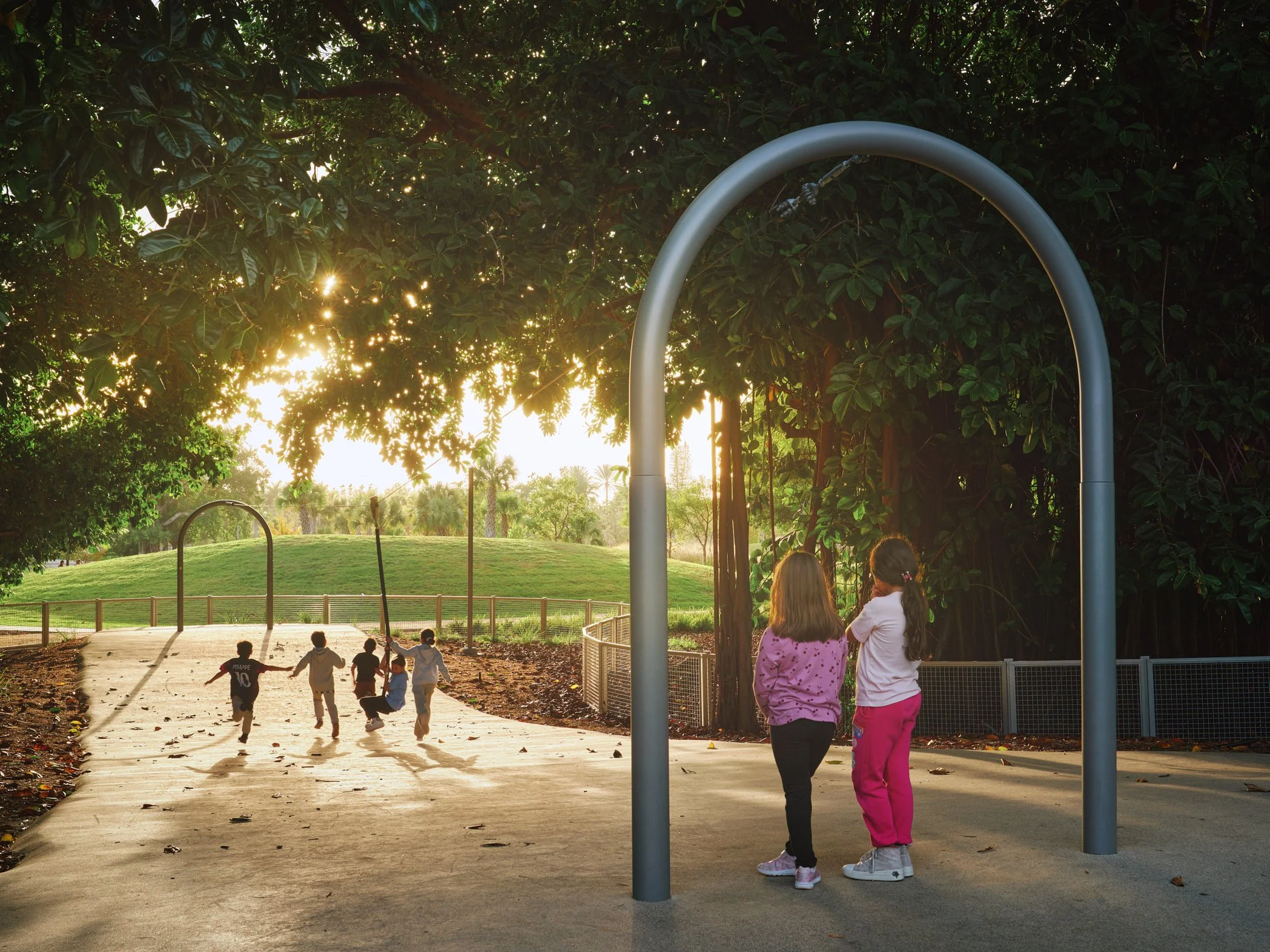 Architectural photograph of children running through a shaded zip-line path beneath a large banyan tree canopy at Bayshore Park in Miami during golden hour. Captured by Miami architectural and travel photographer Christian Santiago.