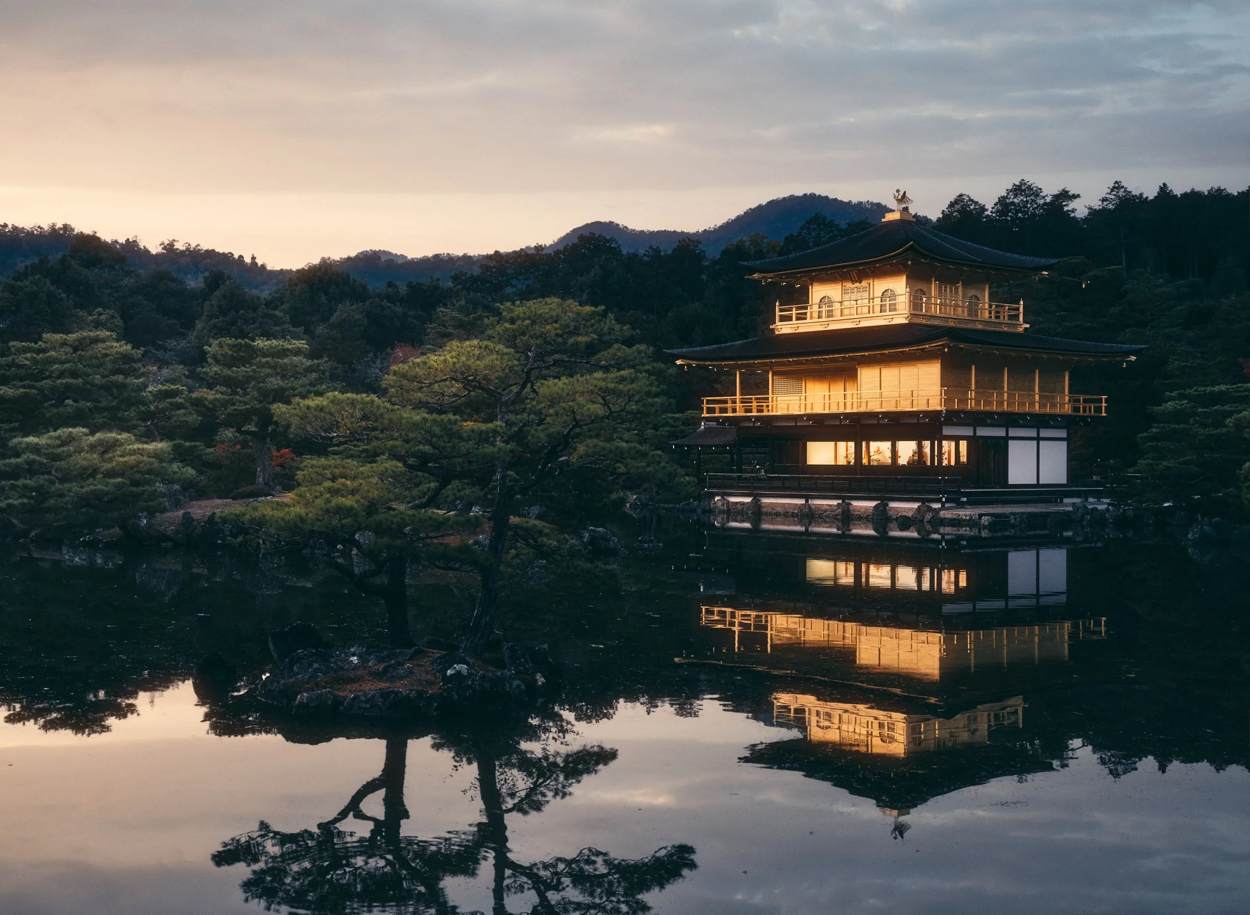 Architectural photograph of Kinkaku-ji, the Golden Pavilion in Kyoto, Japan, captured at dusk. The iconic Zen Buddhist temple glows in warm golden tones, reflecting perfectly in the still waters of the surrounding pond. Framed by lush pines and dista