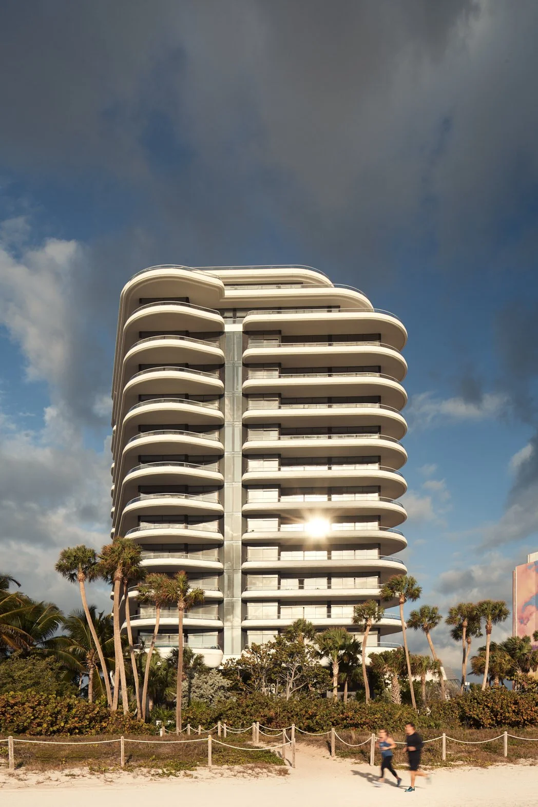 Architectural photograph of Faena House in Miami Beach, a modern luxury residential tower with curving white balconies glowing against dramatic skies at golden hour. The image captures the sleek oceanfront presence of the building with palm trees and