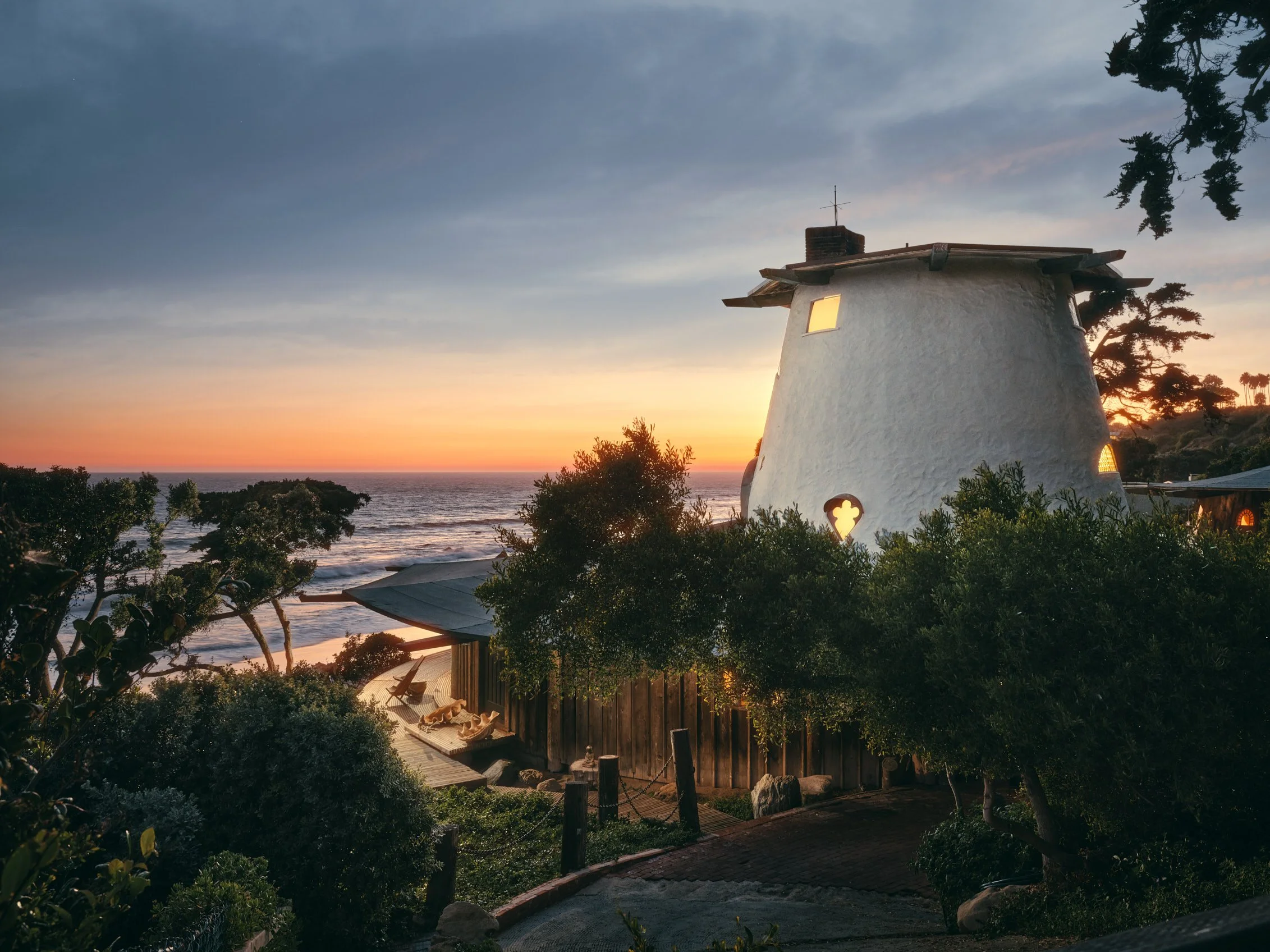 Architectural photograph of the Sandcastle House at sunset, highlighting the iconic whitewashed turret with quatrefoil window and warm interior glow. Perched above the Malibu coastline, the home blends romantic whimsy with organic design, rooted in s