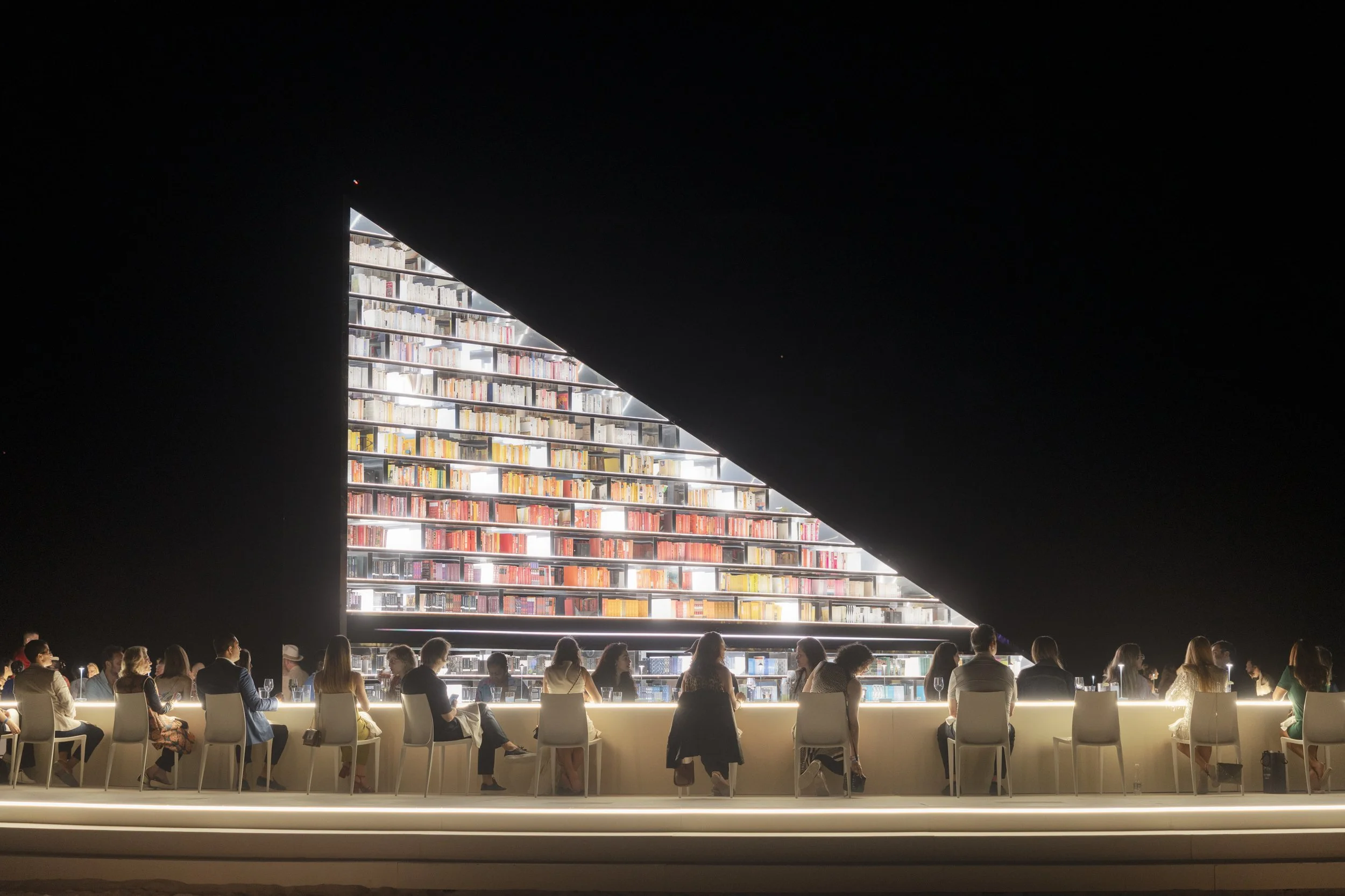 Architectural photograph of Es Devlin’s Library of Us at Art Basel Miami Beach, featuring guests seated around the illuminated installation during a nighttime activation at Faena Miami Beach. The image captures the transformation of the structure int