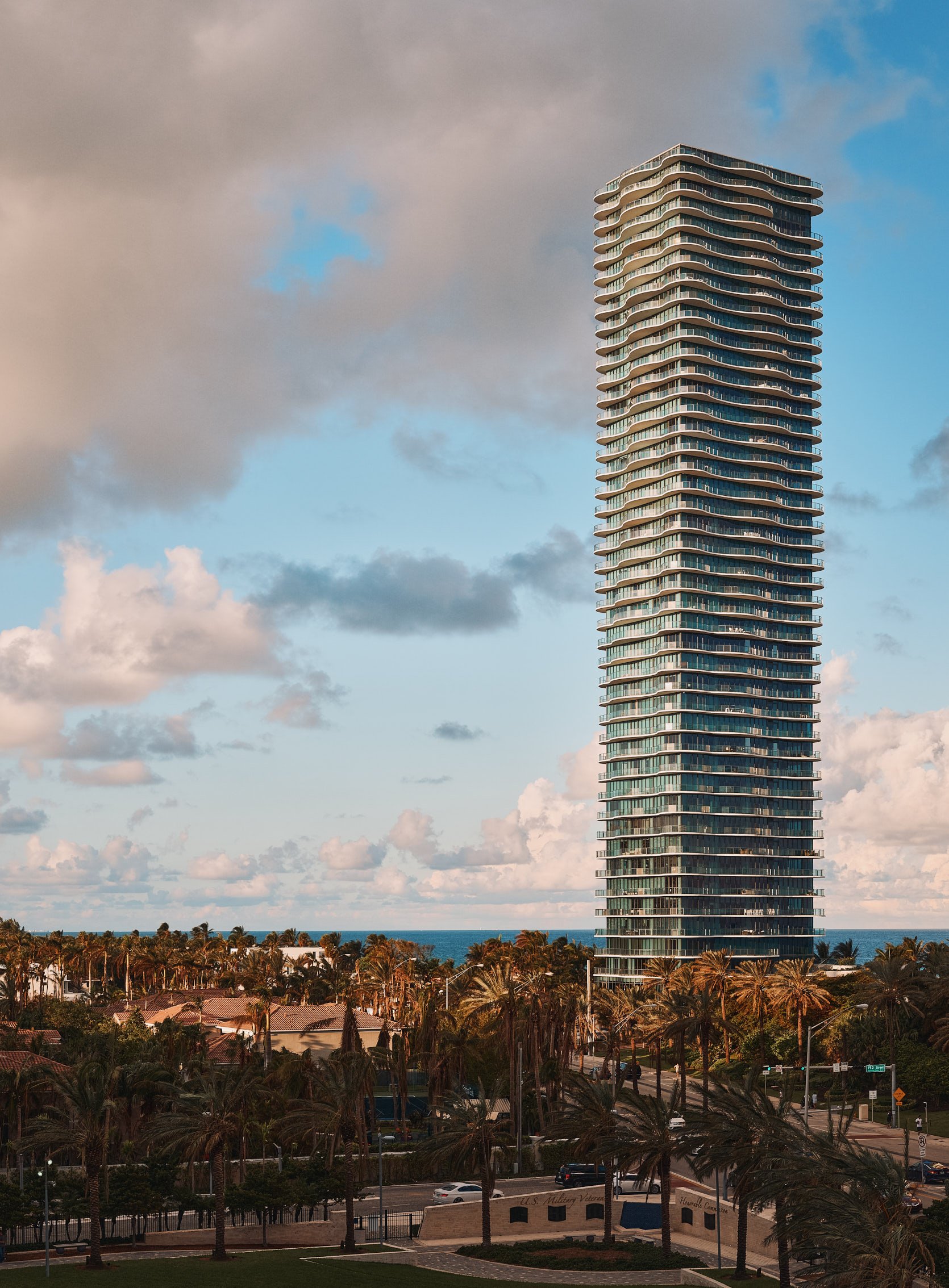 Architectural photograph of Regalia, a luxury oceanfront skyscraper in Sunny Isles Beach, Miami, captured in golden light against a backdrop of palm trees, ocean, and dramatic clouds. The tower’s organic, flowing balconies create a sculptural silhoue