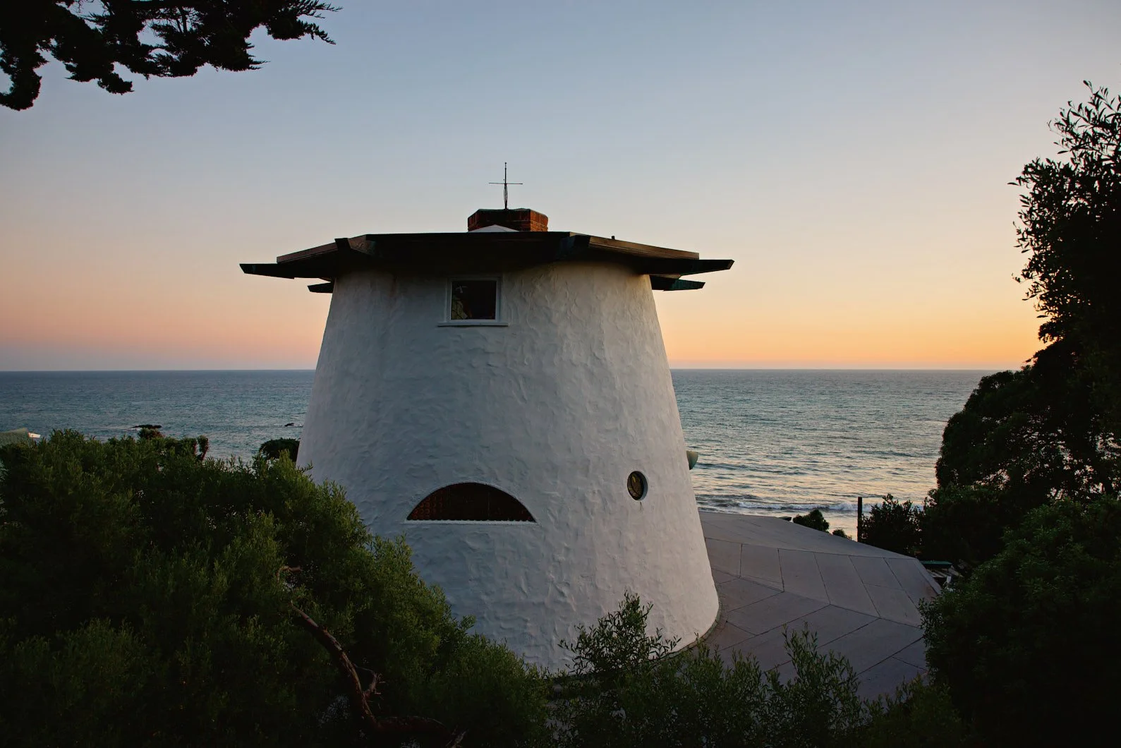 Architectural photograph of the Sandcastle House’s iconic tower, designed by Malibu architect Harry Gesner. Captured at twilight with the Pacific Ocean as a backdrop, the sculptural white structure features porthole windows and a distinctive hand-for