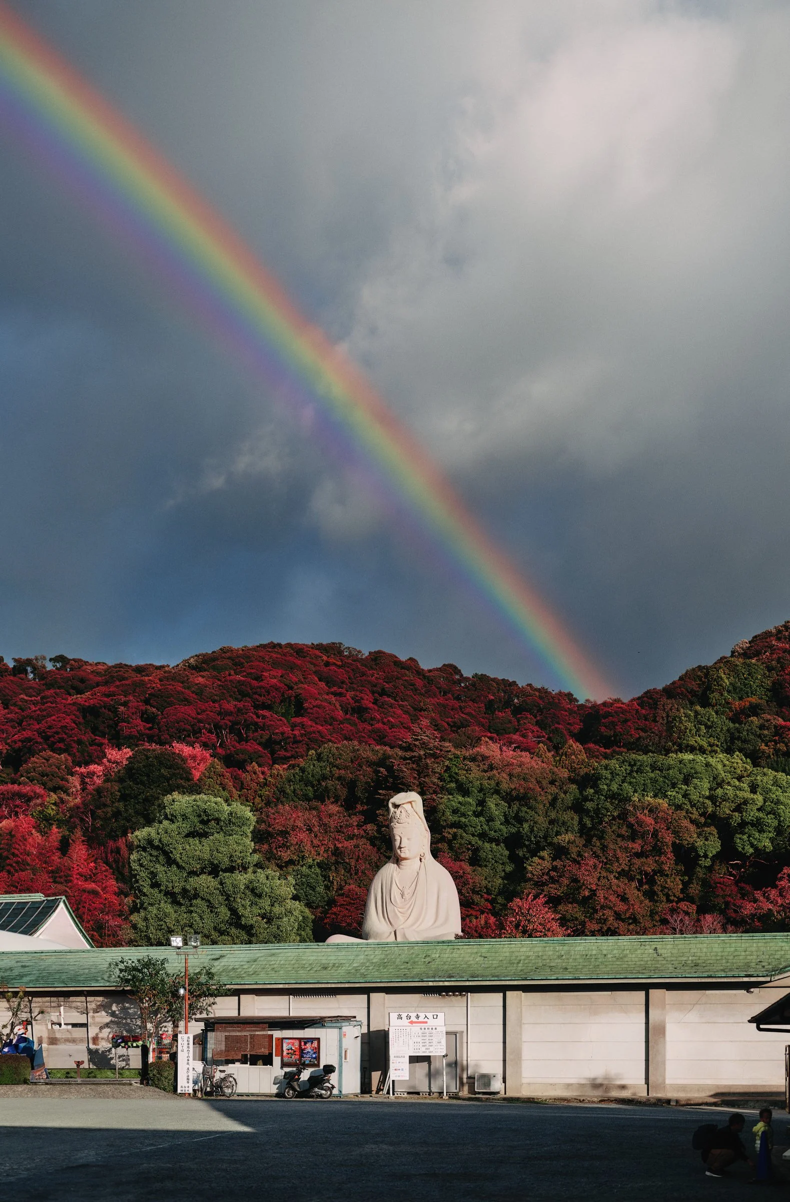 Architectural and cultural photograph of the Ryozen Kannon statue in Kyoto, Japan, framed beneath a vivid rainbow arching over autumn-colored hills. The scene blends striking natural elements with a serene Buddhist monument, evoking both tranquility