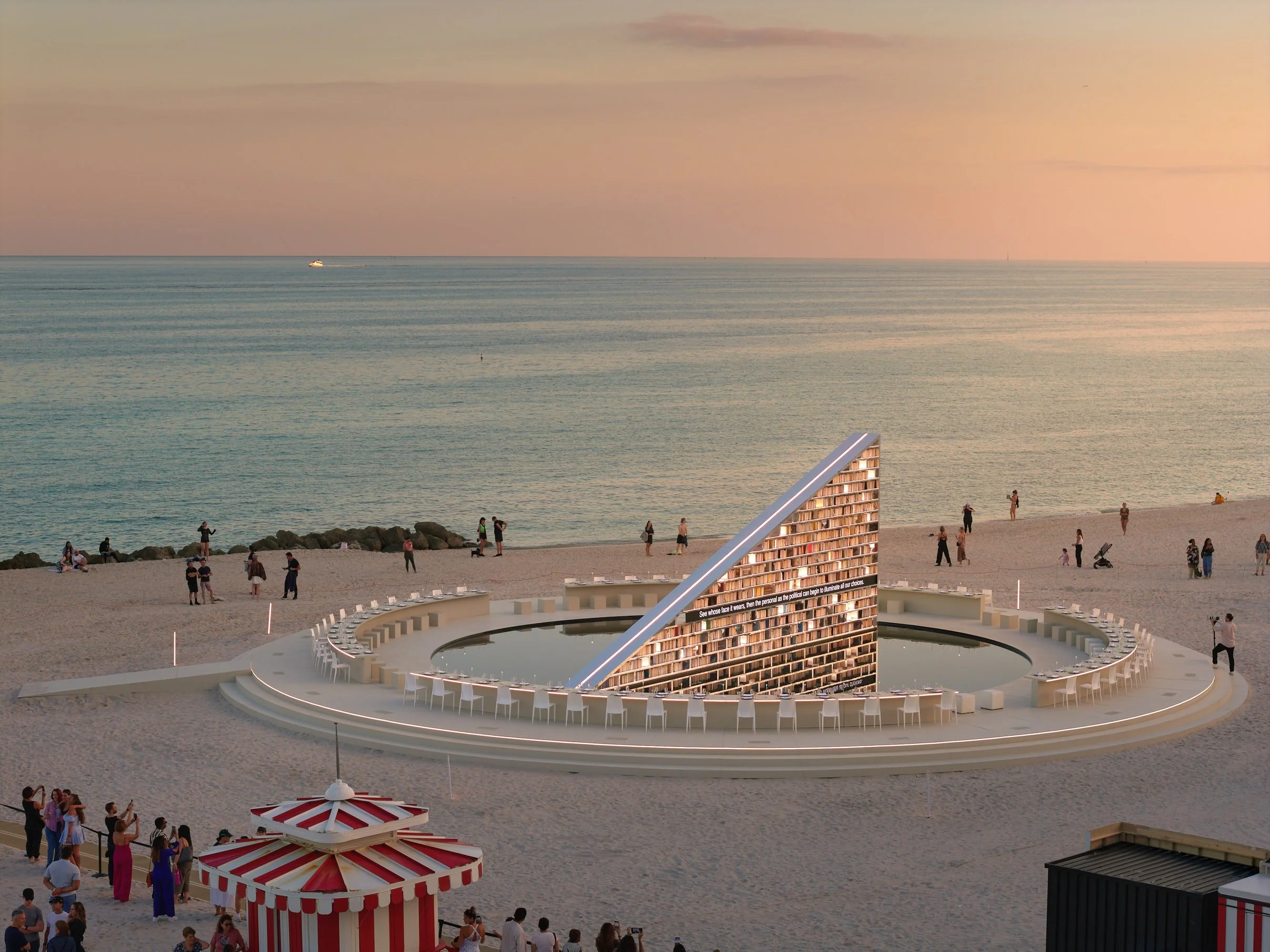 Architectural photograph of Es Devlin’s Library of Us at Art Basel Miami Beach, featuring the illuminated triangular installation set within a circular beachfront plaza at Faena Miami Beach, with visitors engaging around the space during sunset. The 