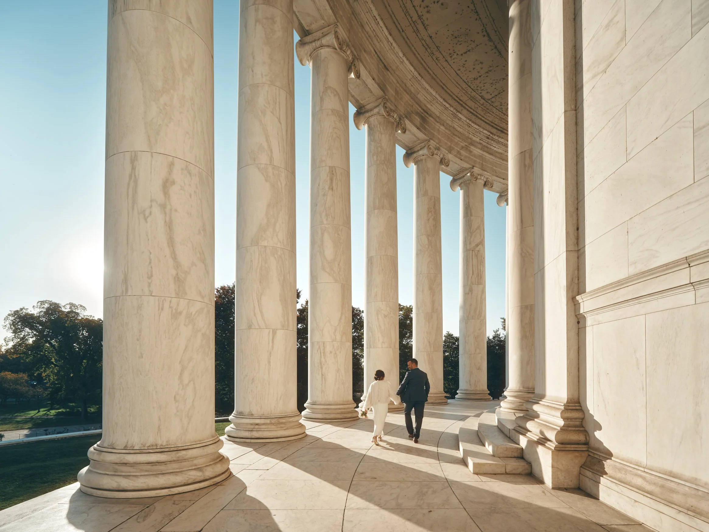 Architectural photograph of the Jefferson Memorial in Washington, D.C., showcasing its neoclassical colonnade bathed in warm golden-hour light. A couple walks hand in hand through the grand marble columns, adding a human element and narrative depth t