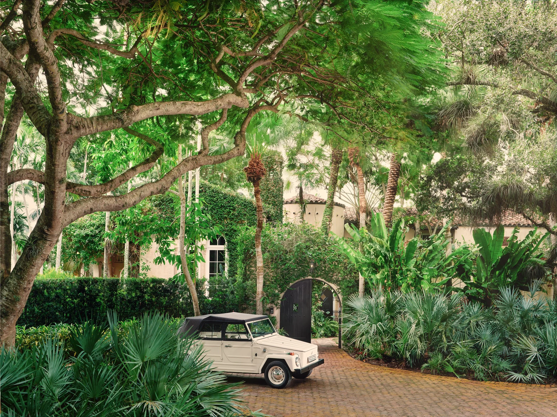 Architectural photograph of a richly layered residential family room interior featuring bold floral wallpaper, a built-in bookshelf flanked by arched doorways, eclectic vintage furnishings in coral, pink, and chartreuse, and a Murano glass ceiling fi
